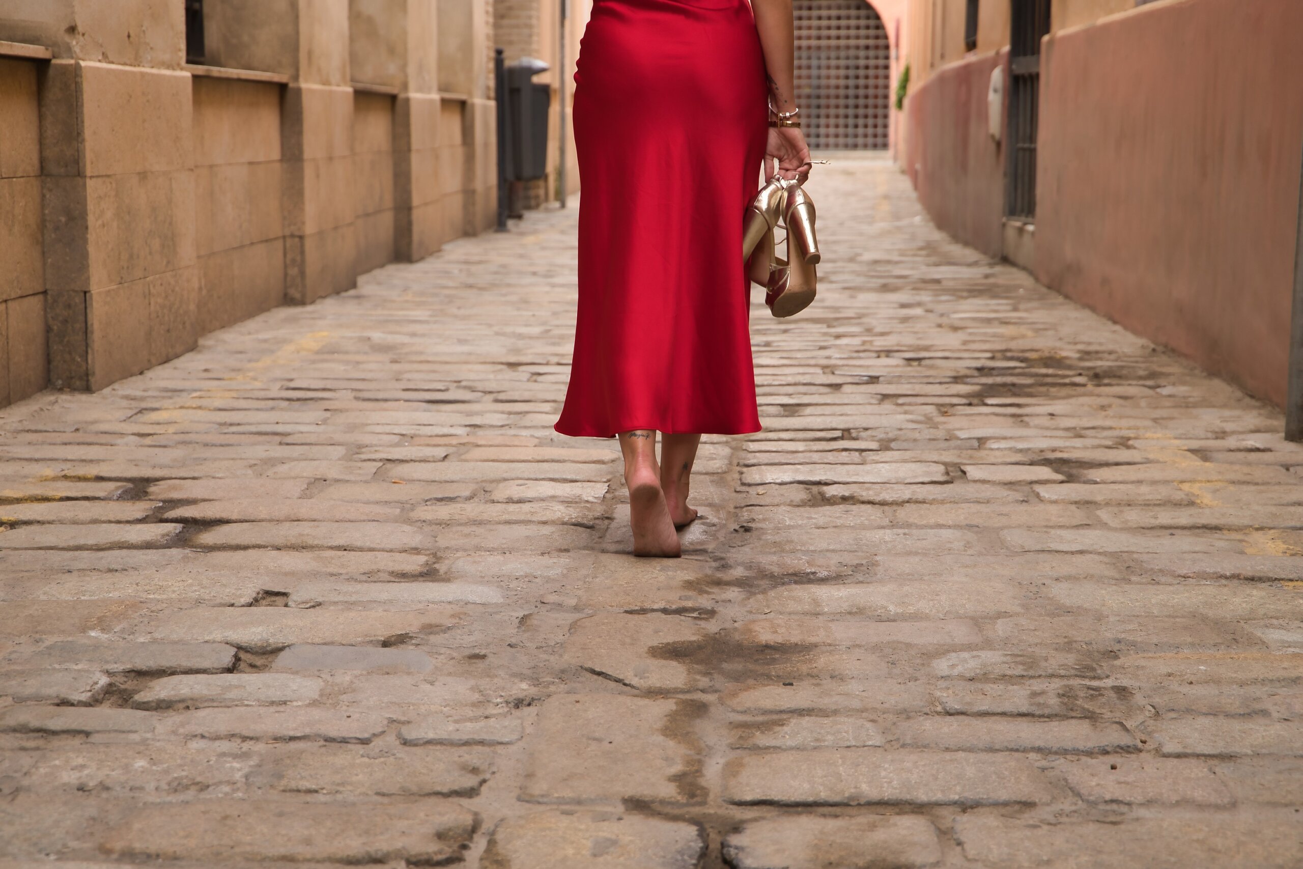 Detail of woman with her back turned, wearing an elegant red party dress and holding golden high heels in her hand, walking barefoot down a city alley.