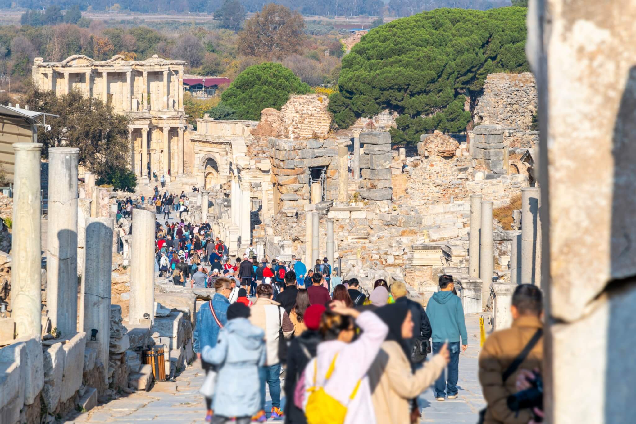 Large tourist crowds walking down Curetes Street in Ephesus, Turkey, with the Celsus Library in the background.