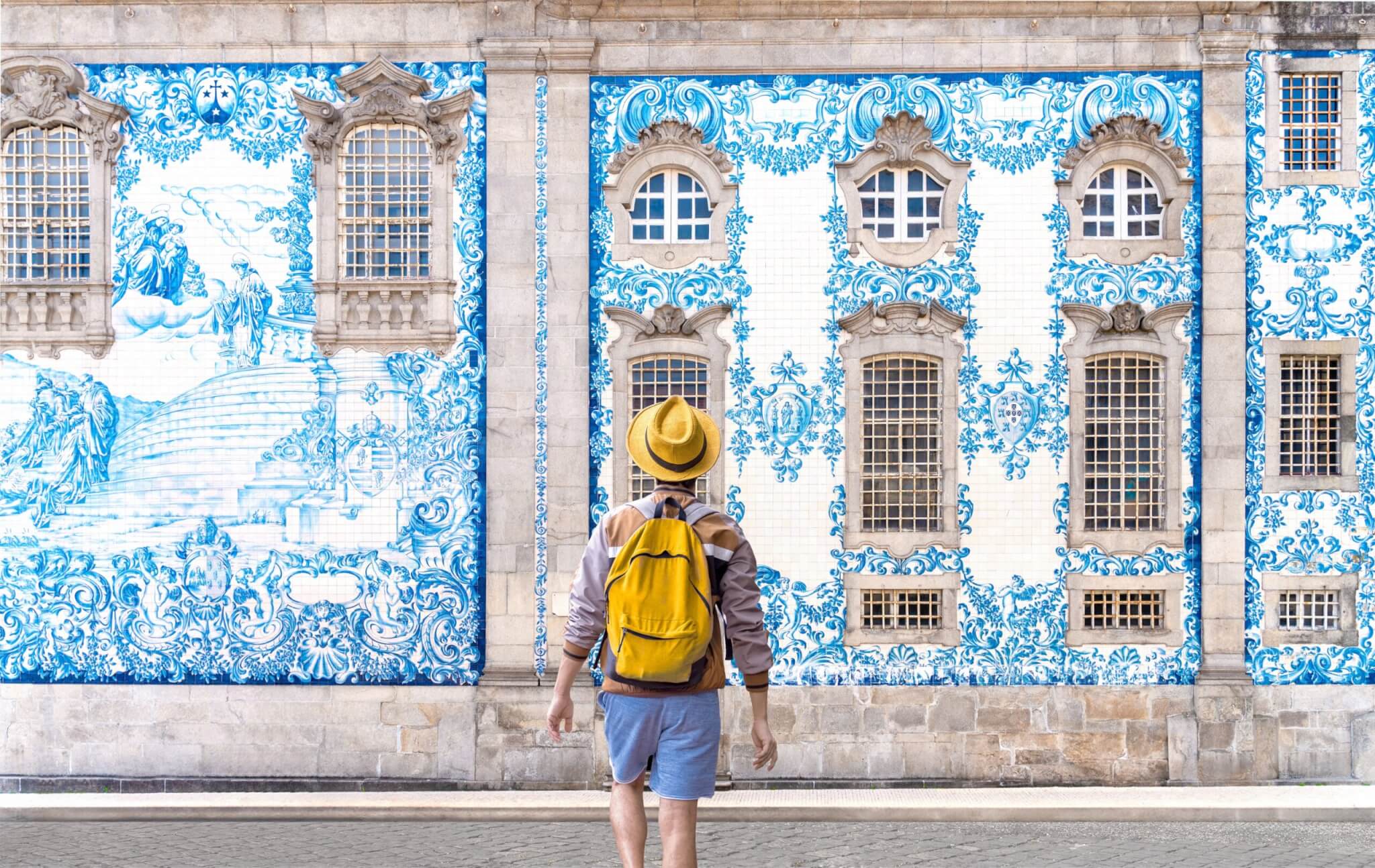 Traveler man with hat and yellow backpack visiting Porto in Portugal - Young tourist enjoy the painted blue tile wall at Carmo church in Clerigos, Porto