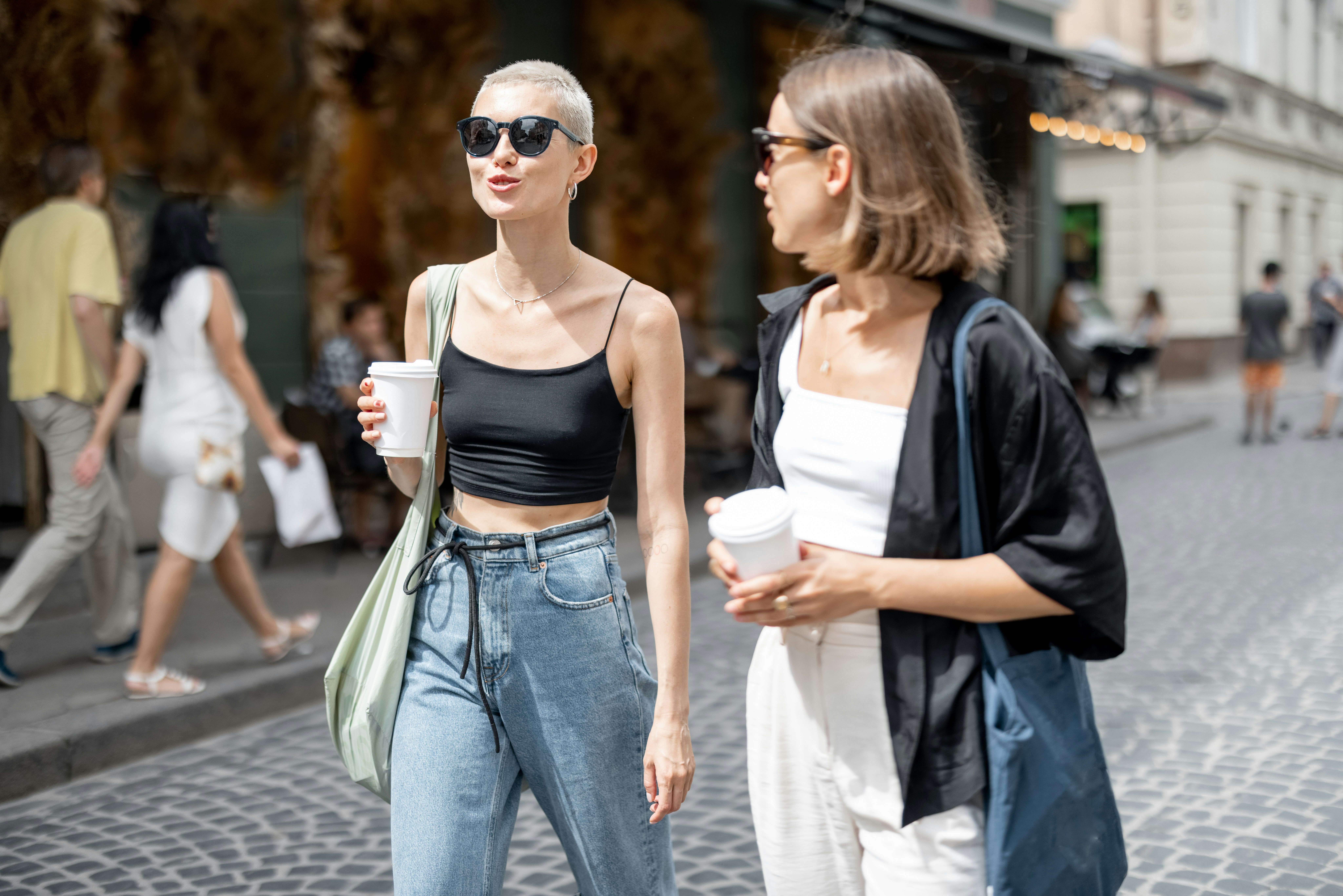 Two women talking on a city street with coffee cups — illustrating why shouting “Is it dangerous around here?” in public can make locals uncomfortable abroad.