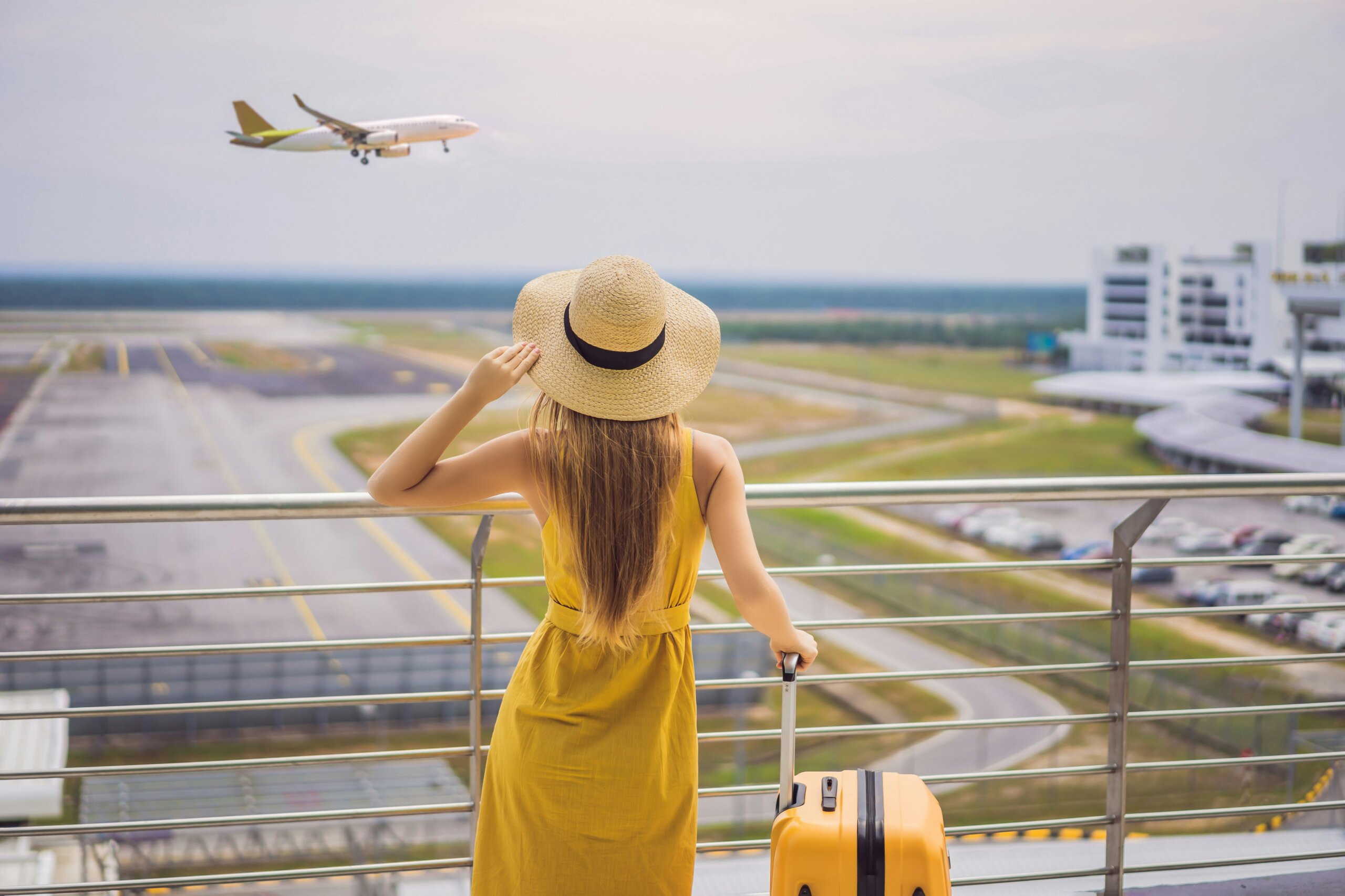 Woman with suitcase watching airplane take off at airport – American traveling abroad concept.