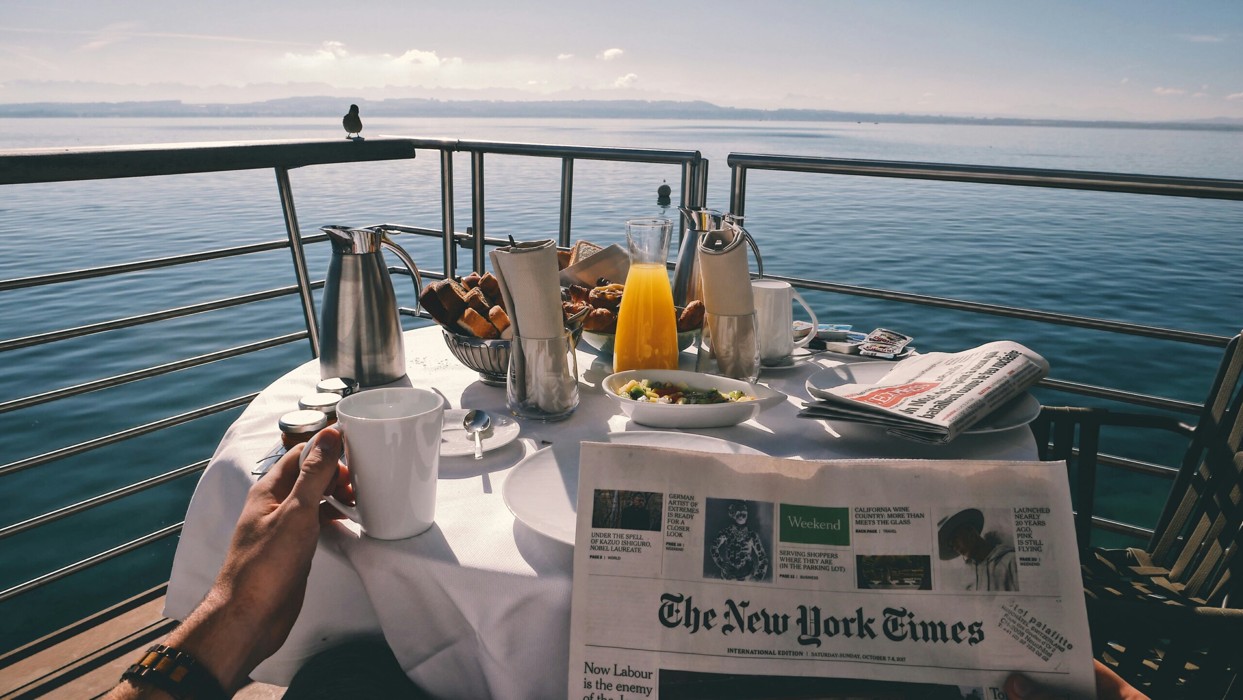 Breakfast table set with food, drinks, and newspaper on a river cruise balcony overlooking calm waters.