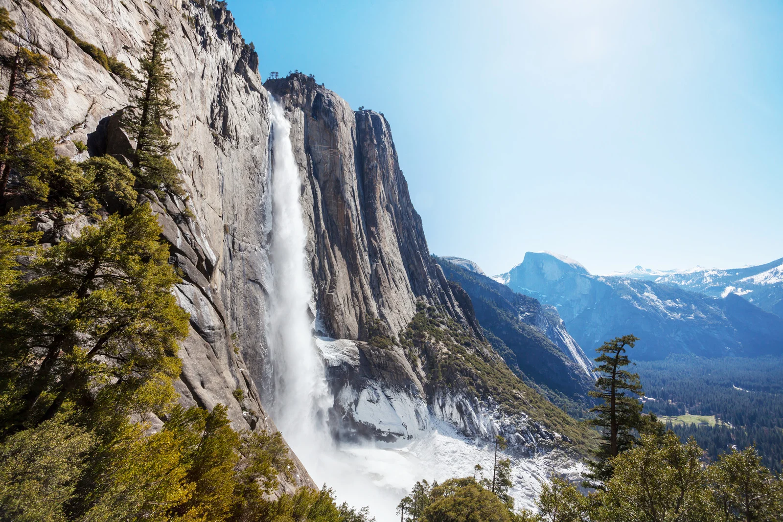 Yosemite Falls cascading down granite cliffs in Yosemite Valley with snowy peaks and forested landscape in the background