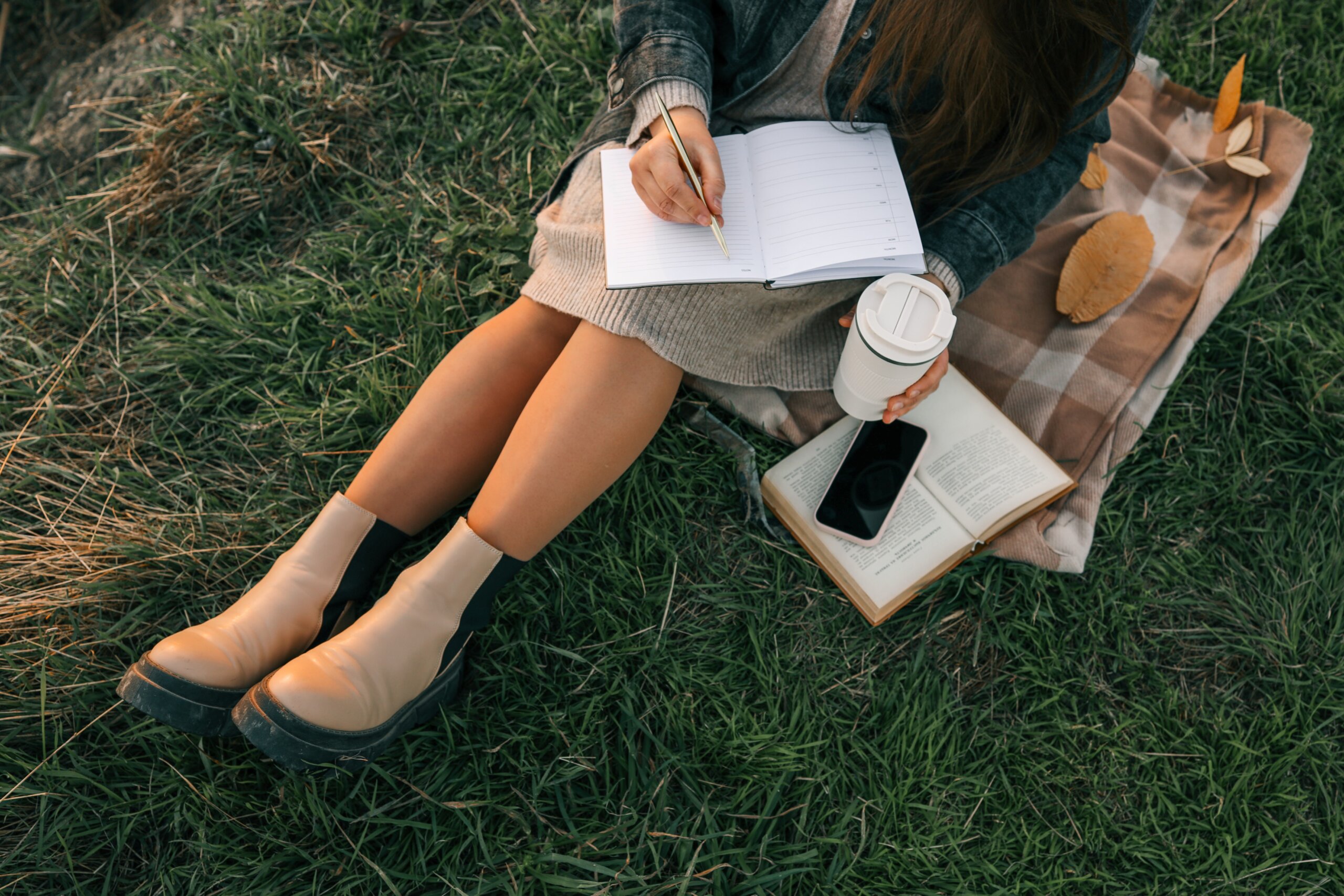 Woman sitting on blanket in grass, writing in a journal with coffee cup in hand. Cozy autumn mood with boots, book, smartphone, and fallen leaves around her
