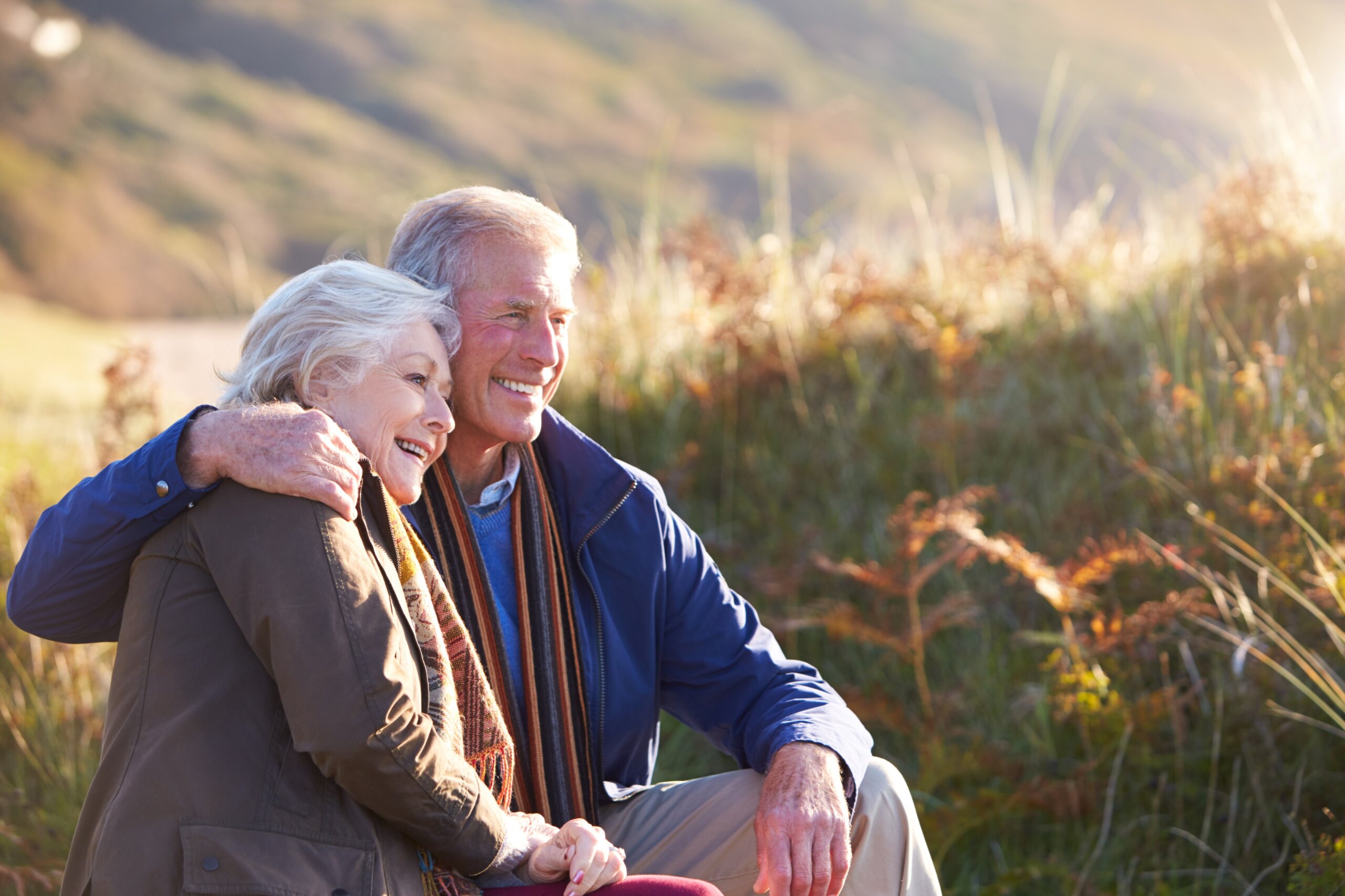 Loving Active Senior Couple Walking Along Coastal Path In Autumn Resting On Rock Together