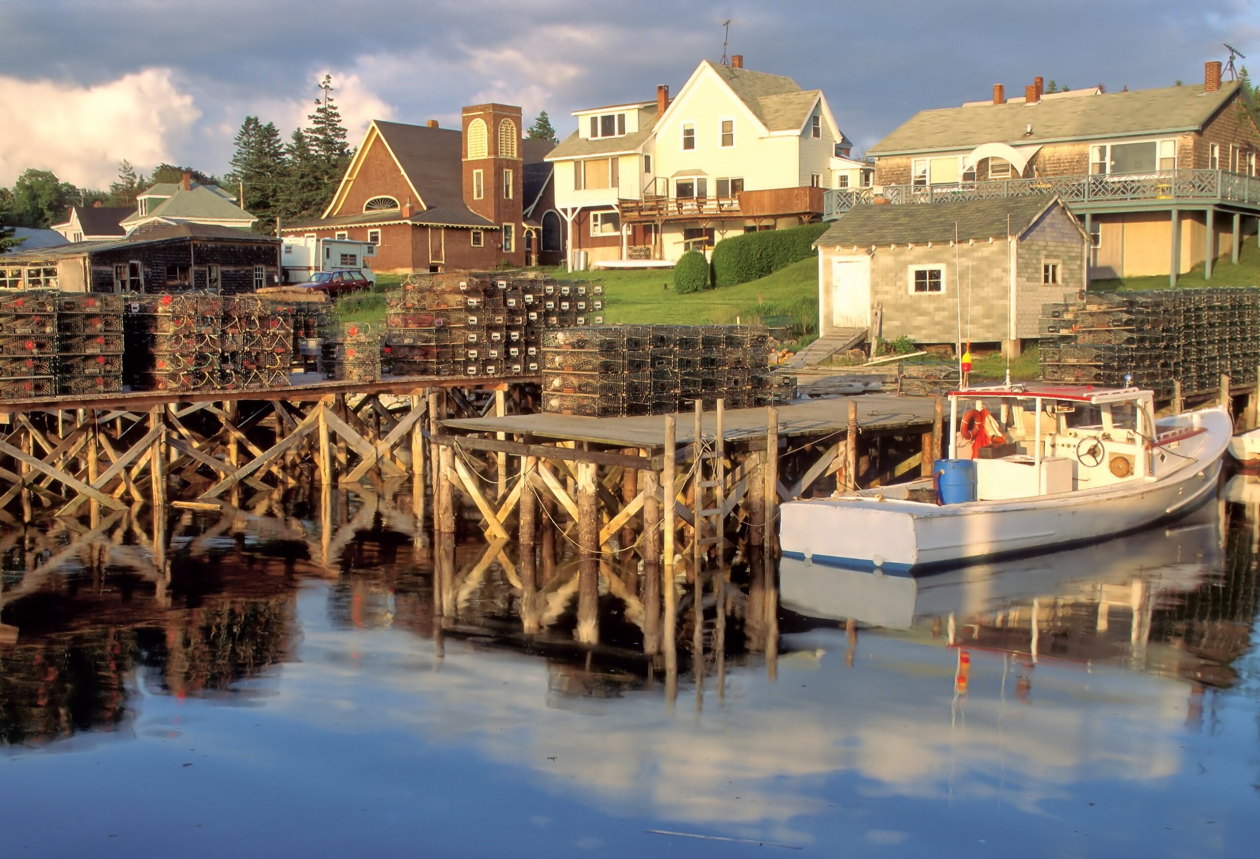 Scenic view of lobster boat and traps, docks, and small church in picturesque coastal fishing village of Port Clyde, Maine on a late summer afternoon with reflections on water.
