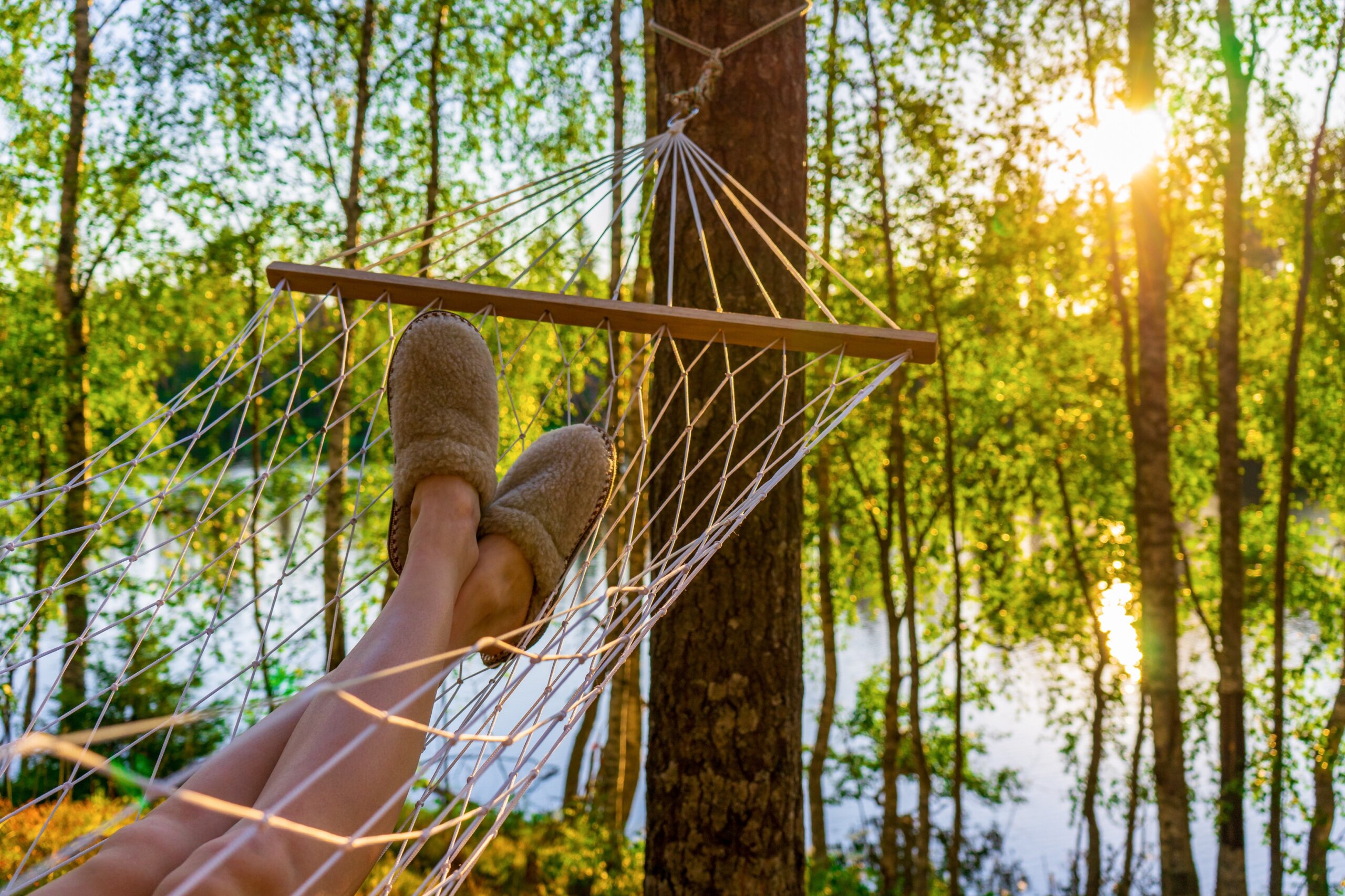Female legs in home slippers in hammock against beautiful summer landscape of northern forest and lake in summer evening at sunset. Leisure, summer vacation, weekend trip, relaxation concept