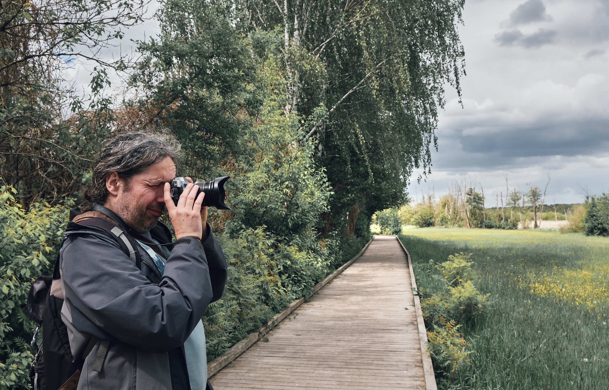 Mature male photographer takes pictures of fields in Germany from wooden walkway. Toned image of fresh green leaves and meadow under grey stormy sky. Local travel, staycation concept