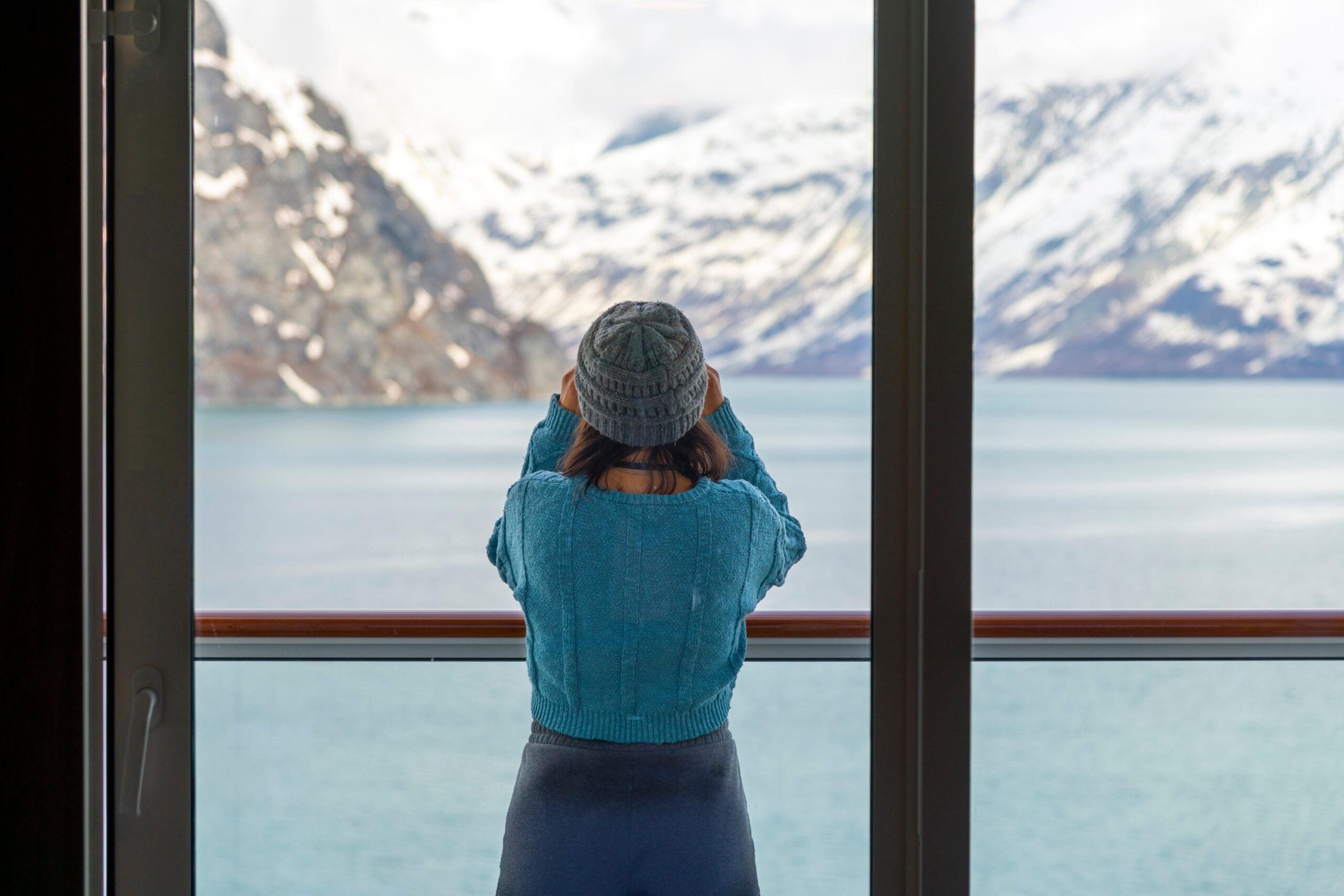 A young woman with binoculars views the snow covered mountains and glaciers from a cruise ship balcony at Glacier Bay National Park and Reserve, Alaska USA