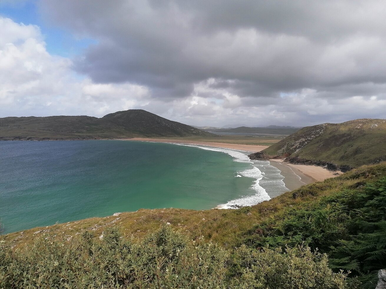 Panoramic view of Trá Mór beach in Donegal, Ireland, showing turquoise waters, a sandy shoreline, and rolling green hills under a cloudy sky.