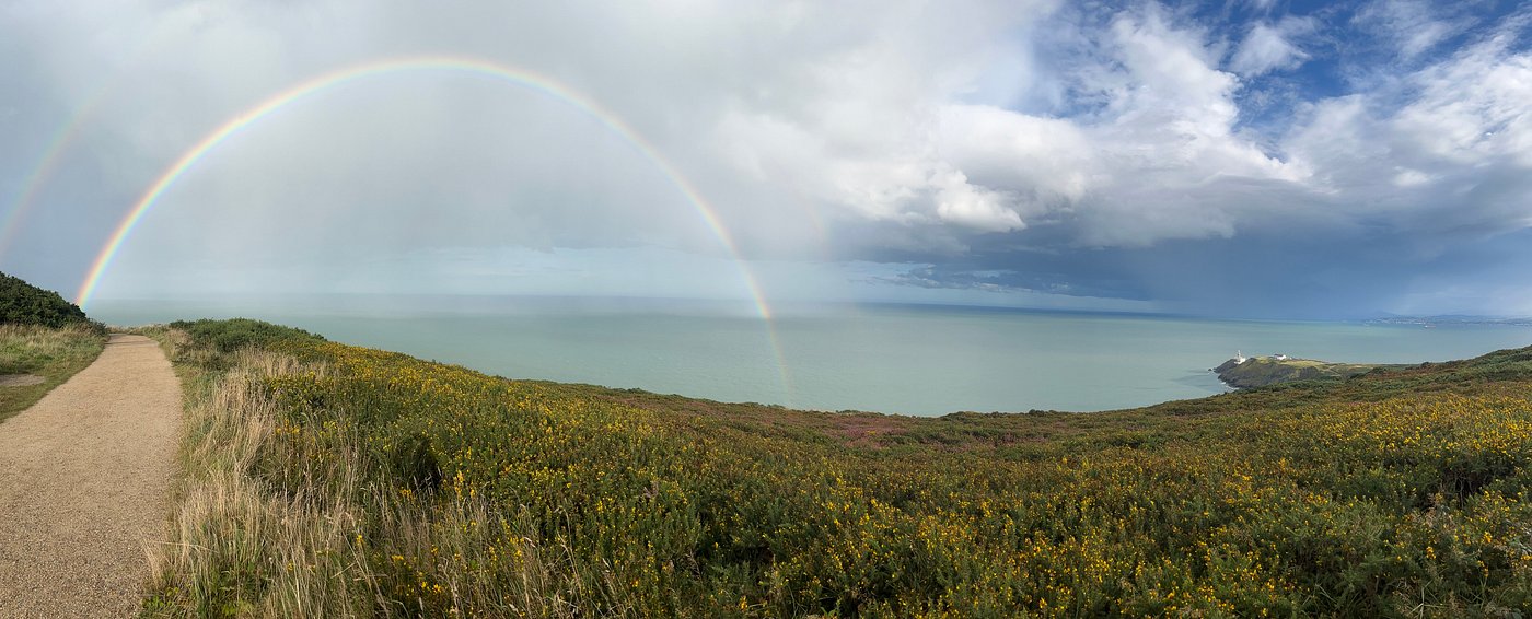 Rainbow arching over the cliffs and coastal path leading to Secret Beach in Howth, Dublin, with the sea and distant headlands in view.