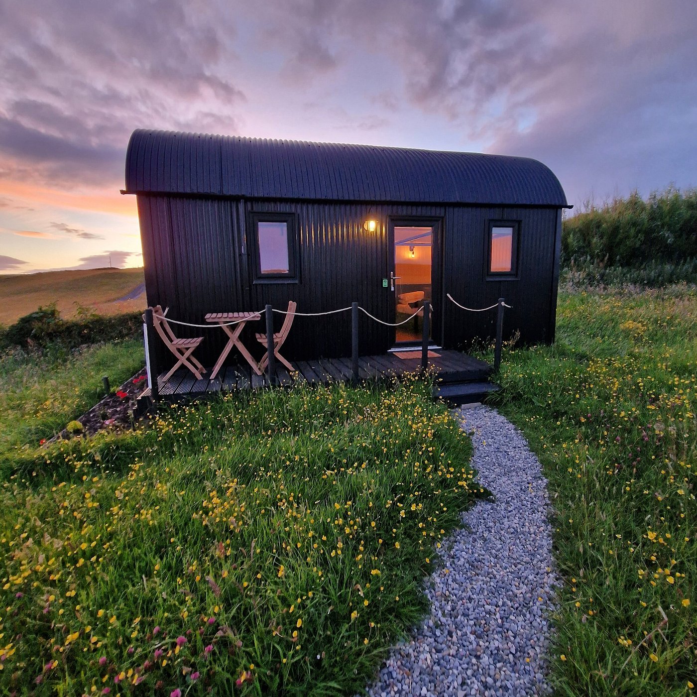 Black shepherd’s hut surrounded by wildflowers with a sunset sky at Wild Meadow Huts in Doolin, County Clare, Ireland.