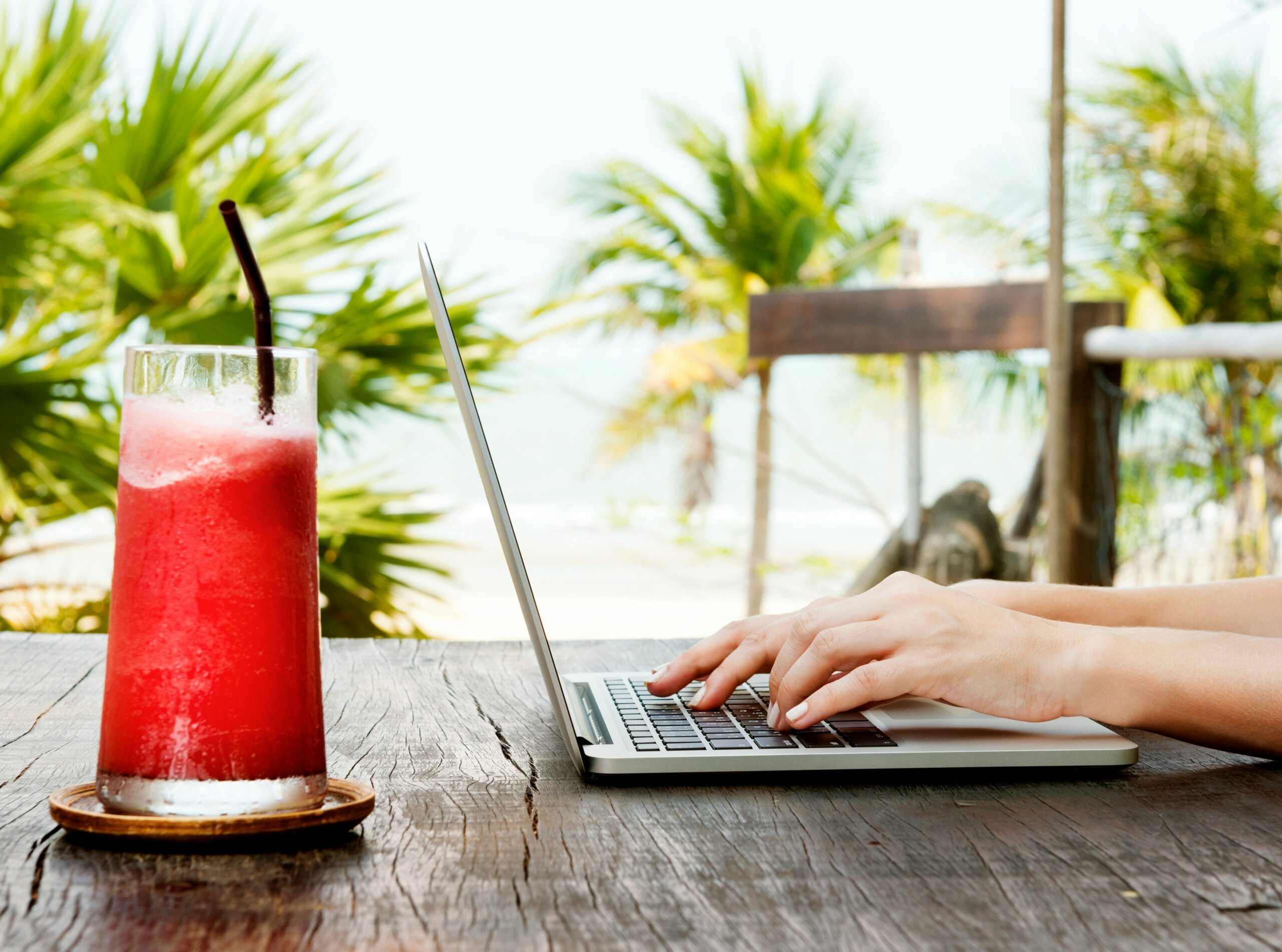 Hands typing on a laptop at a tropical café with a cocktail, depicting the remote work and travel lifestyle.
