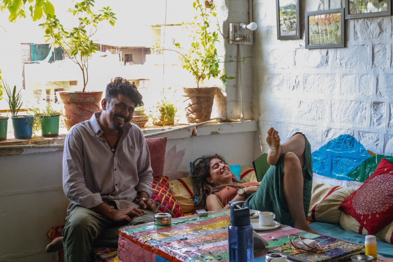 Travelers sitting in colorful Indian hostel sharing tea and conversation surrounded by plants and sunlight.