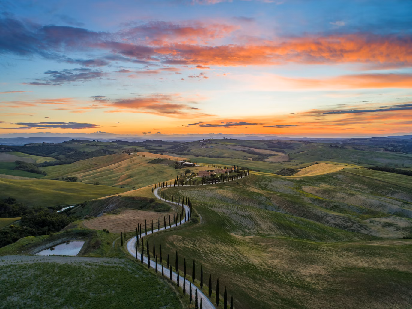 road between green grass field near mountains under blue and brown sky at golden hour - San Quirico d'Orcia, Italy
