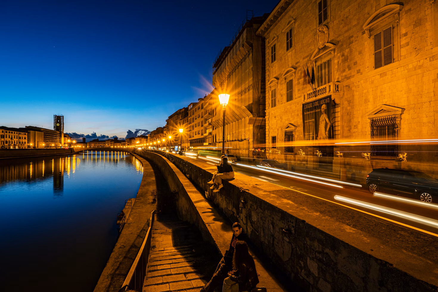 a person sitting on a bench next to a river at night - Pisa, Province of Pisa, Italy
