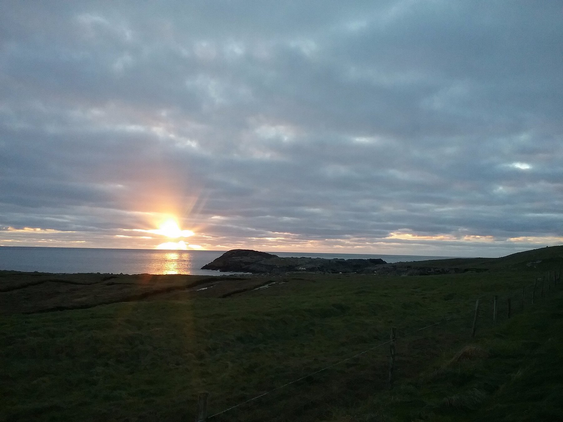 Sunset over Annagh Bay on Achill Island, County Mayo, with the sun breaking through clouds and reflecting on calm sea waters.