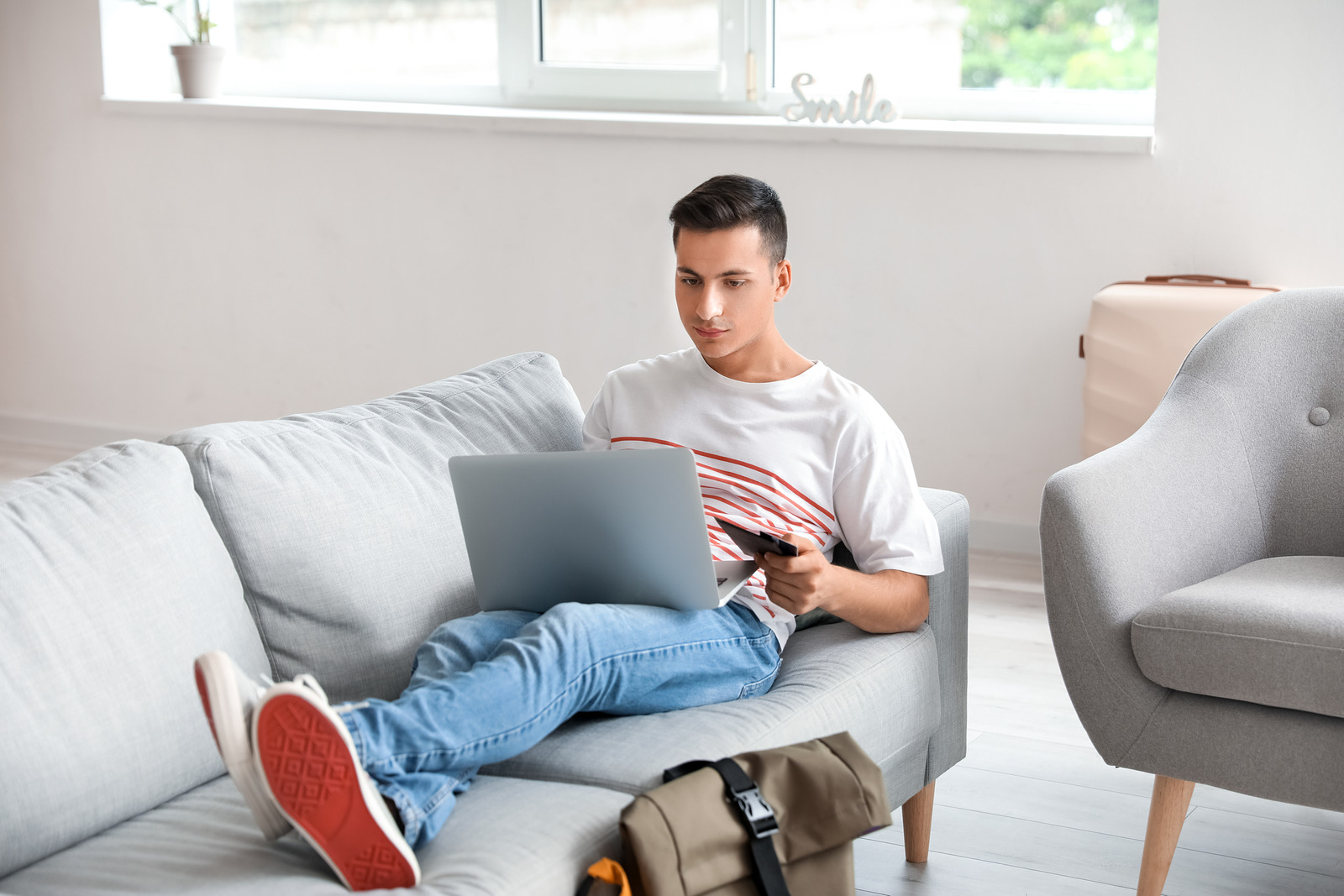 Young man using a laptop and smartphone at home to find cheap flight deals, showcasing budget travel strategies.