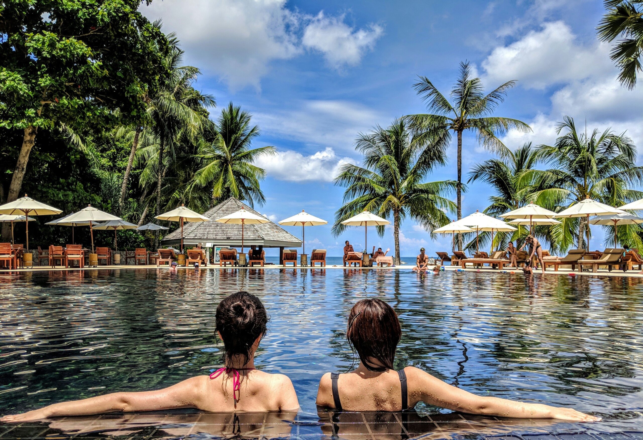 Two women relaxing by a resort pool surrounded by palm trees, inspired by social media travel trends.