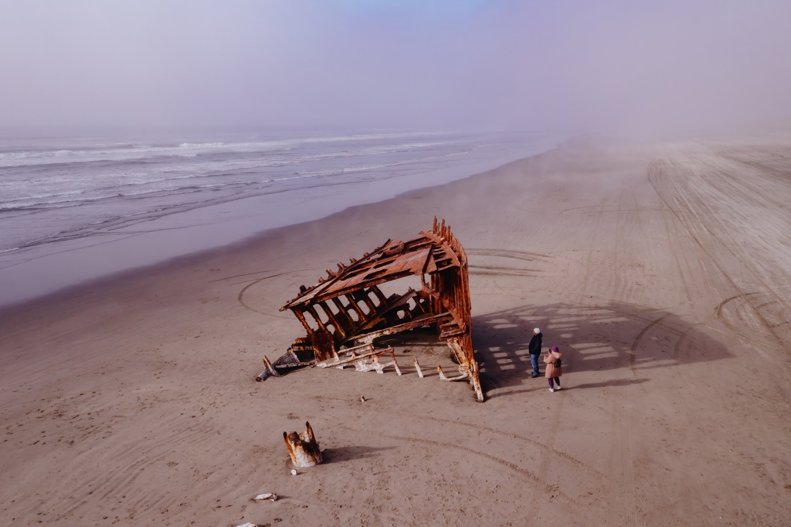 Rusty ship wreck on foggy beach; couple observes the decaying vessel. Coastal scene. Wreck Of The Peter Iredale, Fort Stevens State Park, Oregon, USA