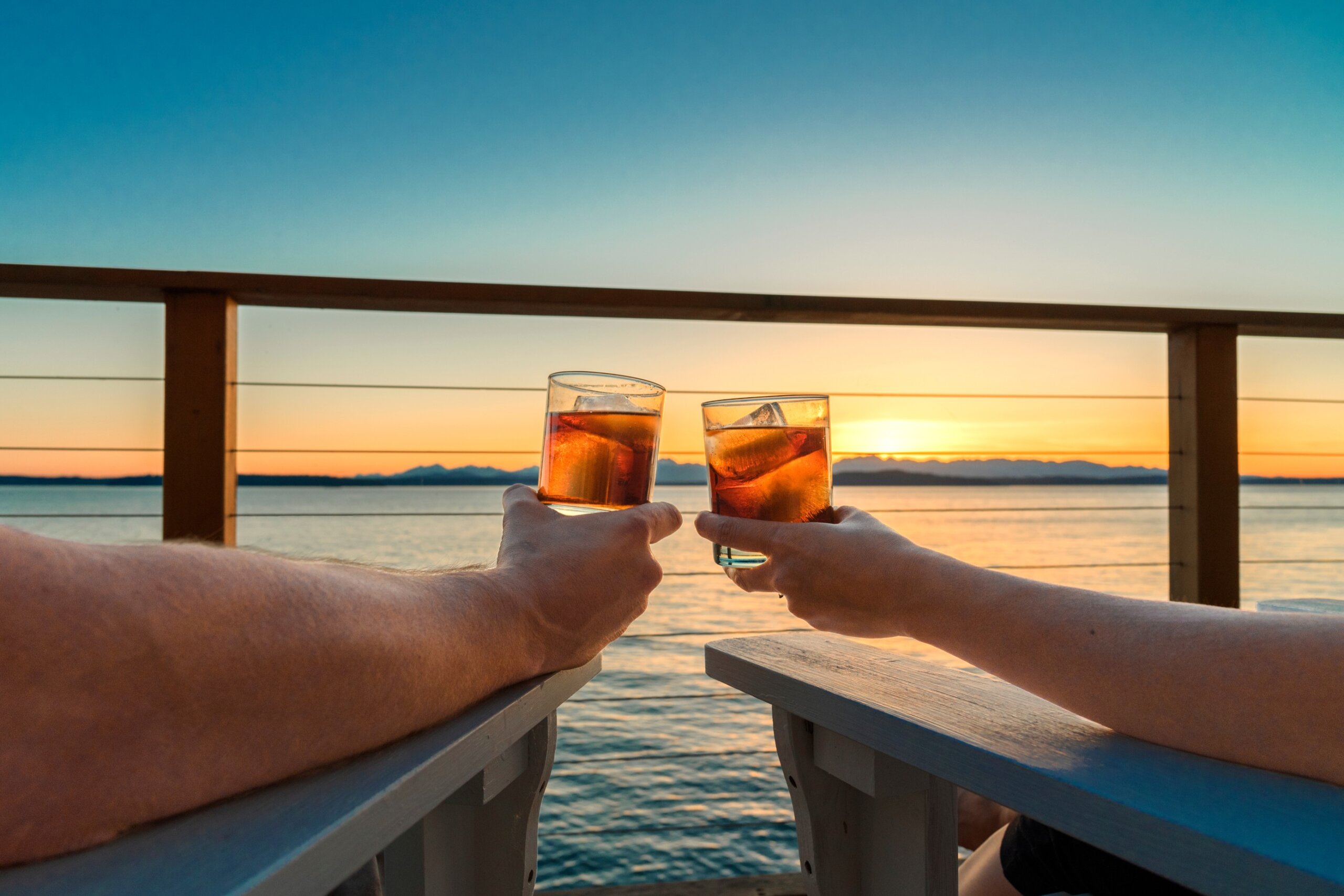Couple holding up icy cocktail drinks while sitting on a seaside deck at sunset.

