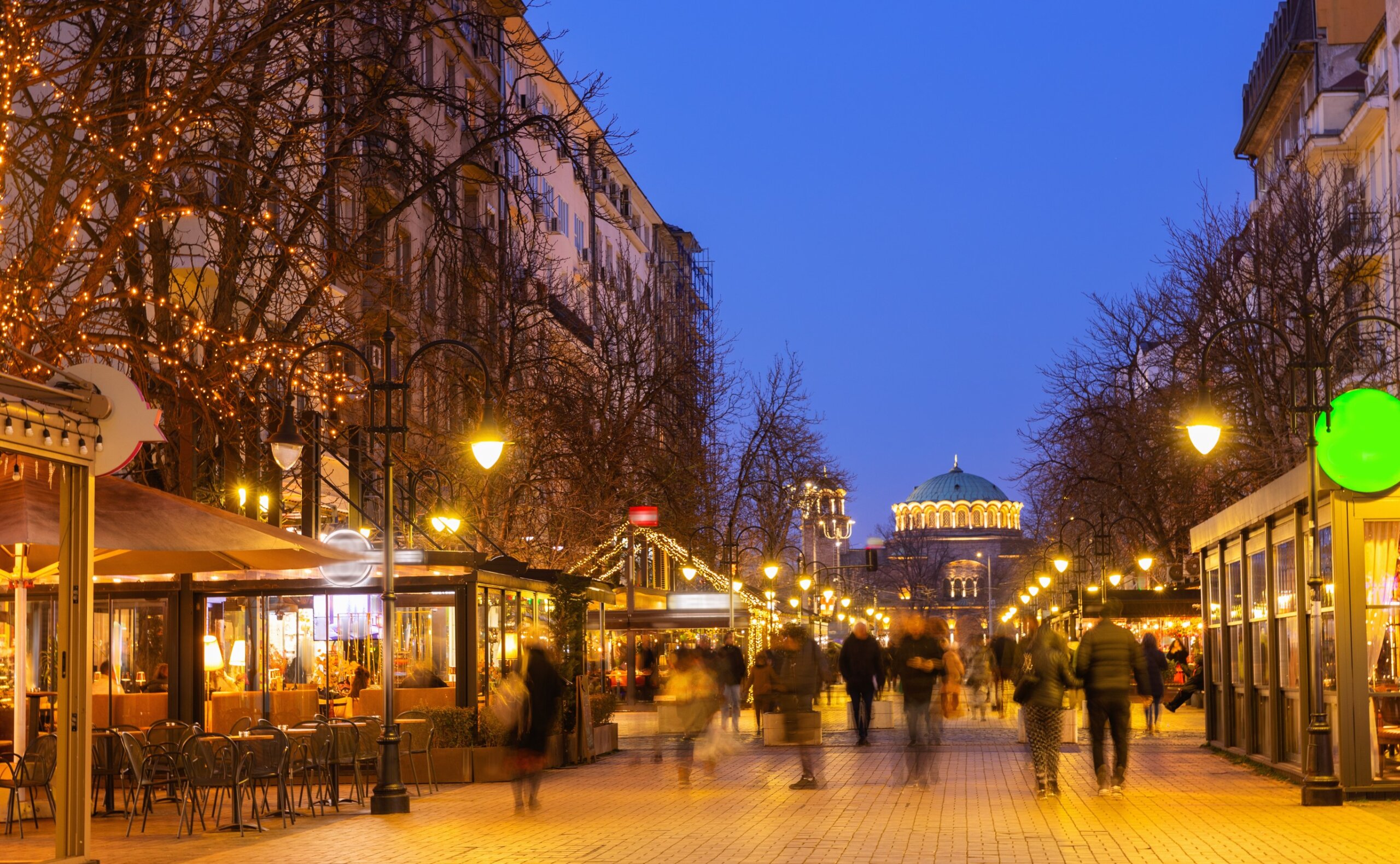 Lively evening scene on Vitosha Boulevard in Sofia, Bulgaria, with blurred pedestrians, glowing streetlights, decorated with festive string lights cafes and lighted St. Nedelya Church in background