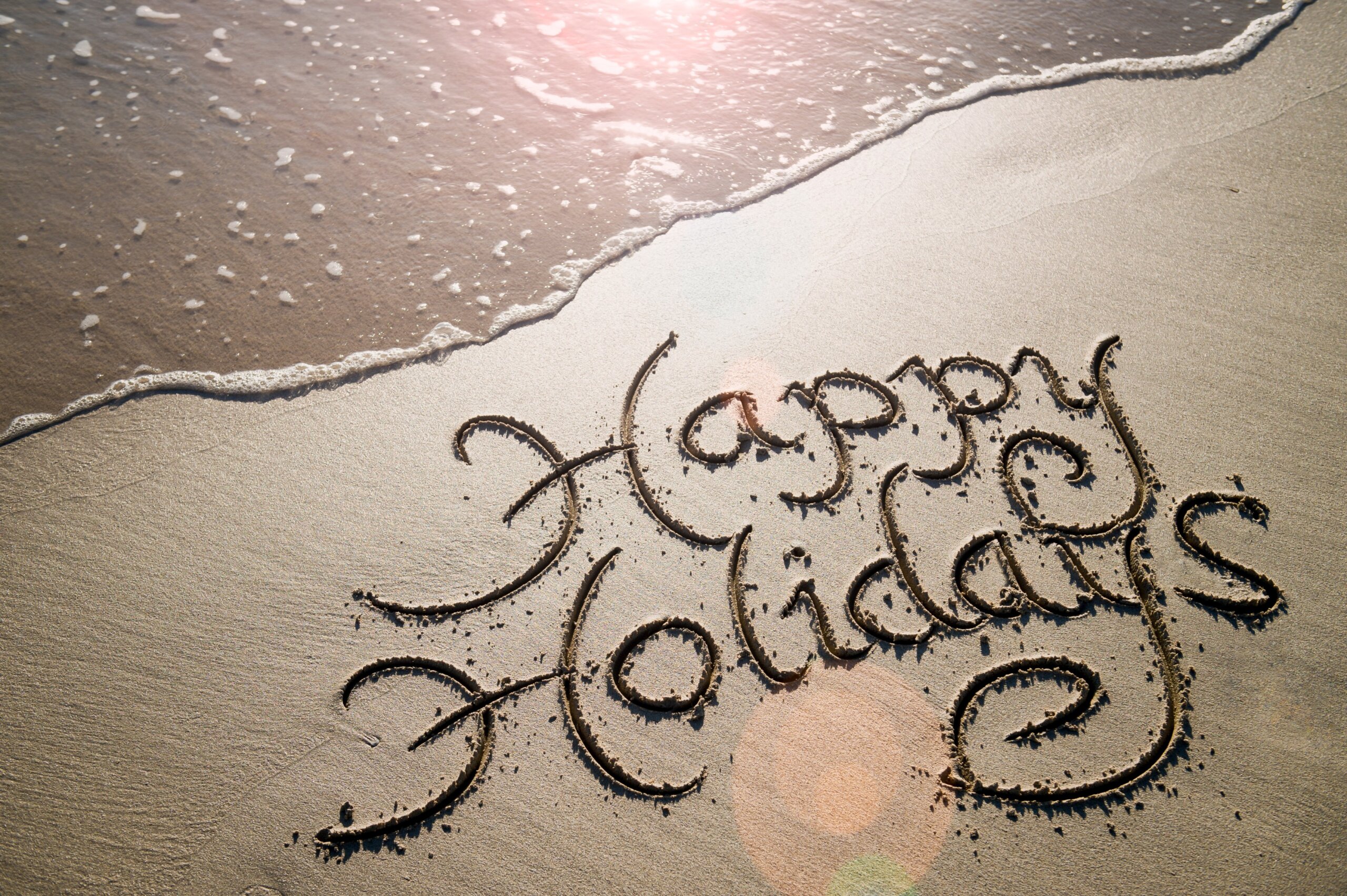 Happy Holidays message handwritten in smooth sand with an oncoming wave in the lens flare of the tropical sun