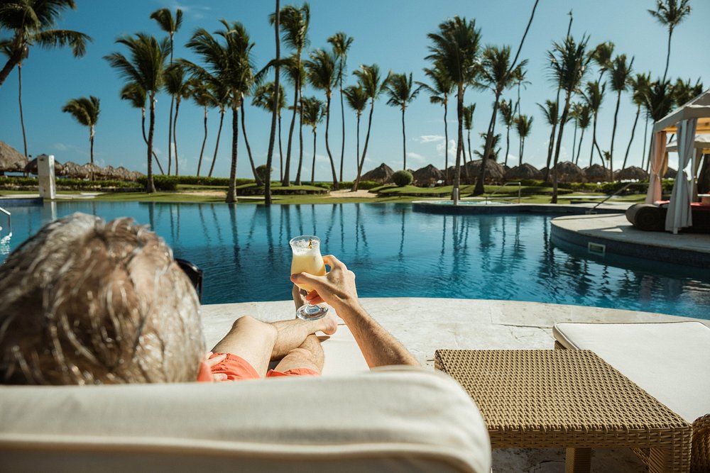 Man relaxing by a tropical resort pool holding a drink with palm trees and ocean in background