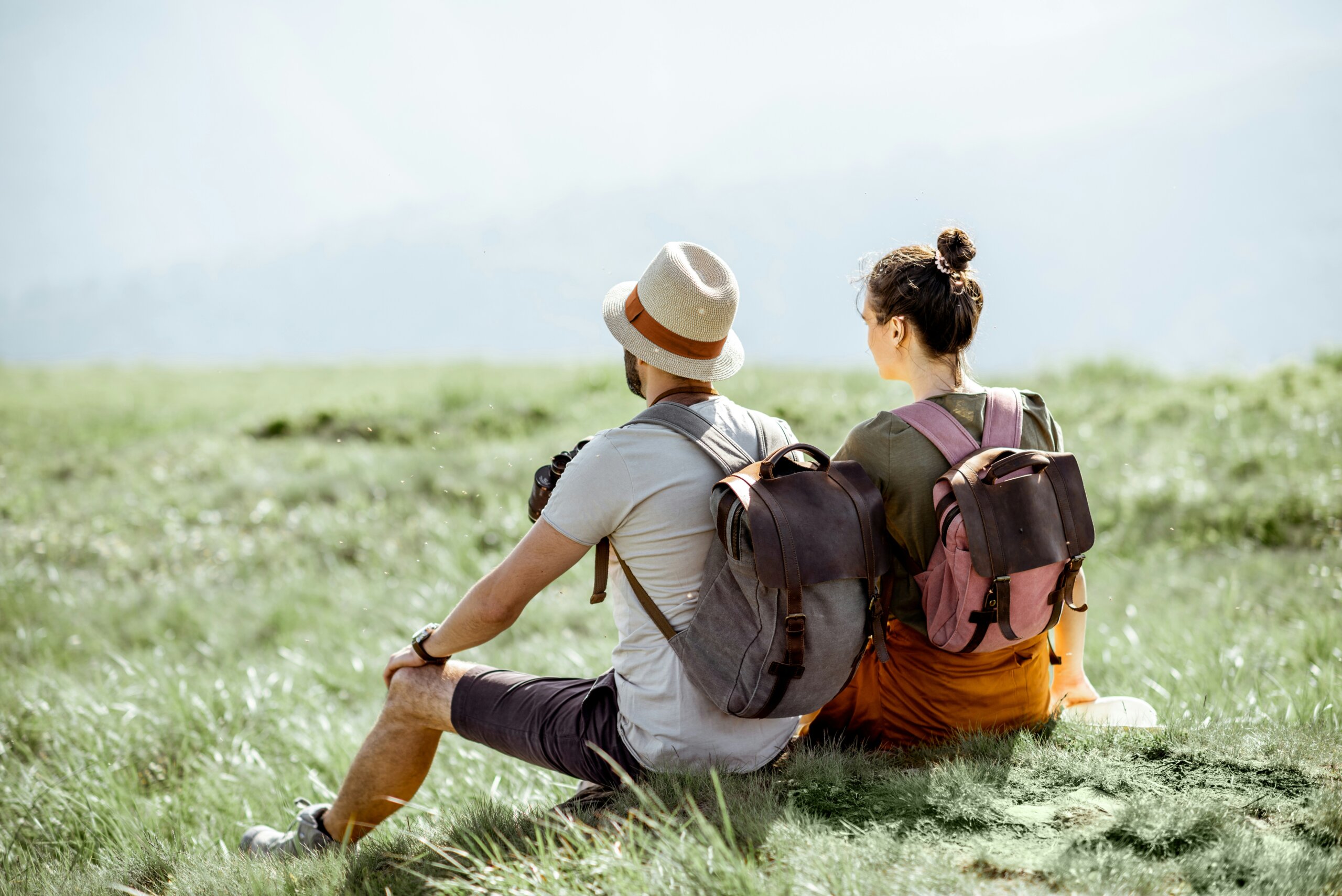 Couple sitting on a grassy hill with backpacks overlooking a scenic mountain landscape during a peaceful outdoor travel moment.