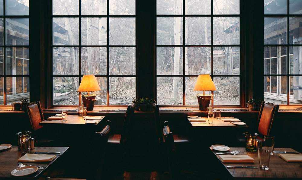 Cozy mountain lodge restaurant interior with warm lighting and snowy forest view through large windows.