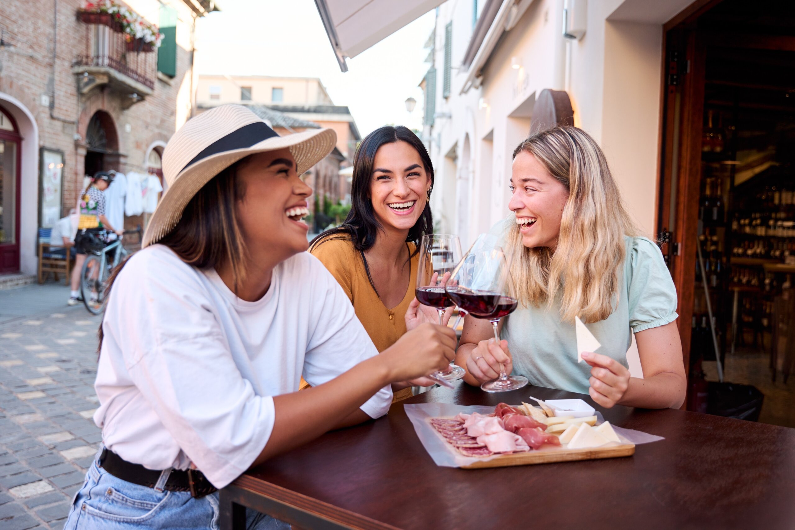 Three happy female friends are toasting with glasses of red wine while sitting at a table outside a restaurant in a picturesque Italian town, enjoying a selection of cheese and cured meats
