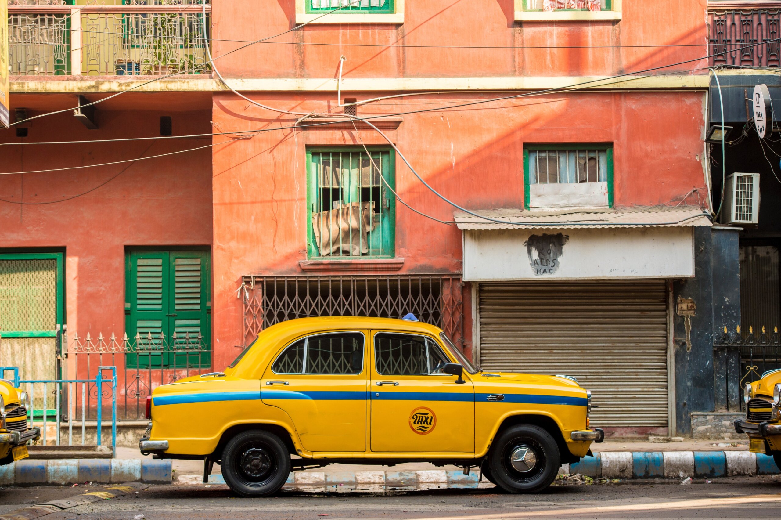 An Ambassador yellow cab taxi is parked in the street under a red-colored house in Kolcata, India
