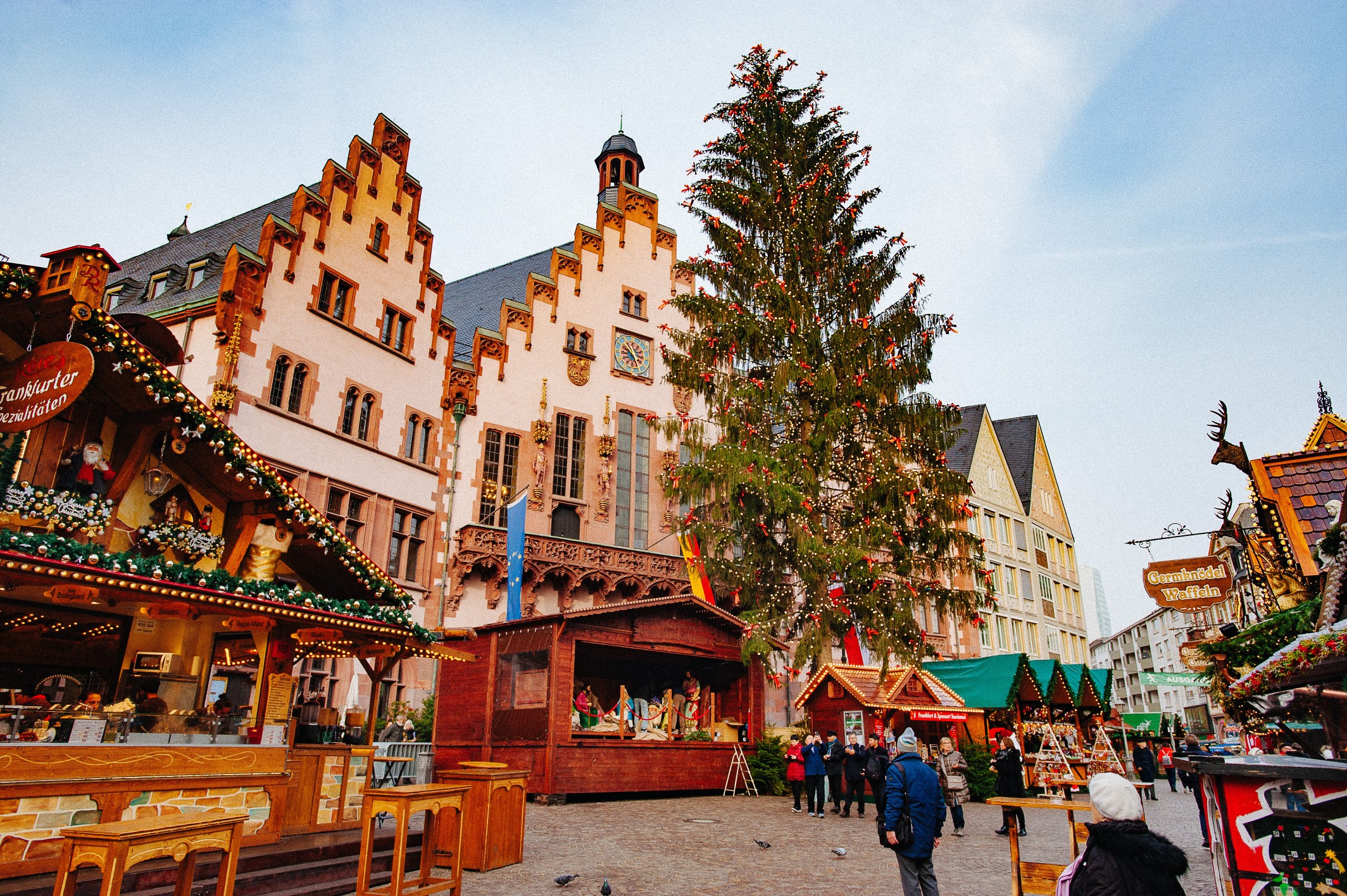 European Christmas market with wooden stalls, holiday lights, and a large decorated tree in a historic town square.