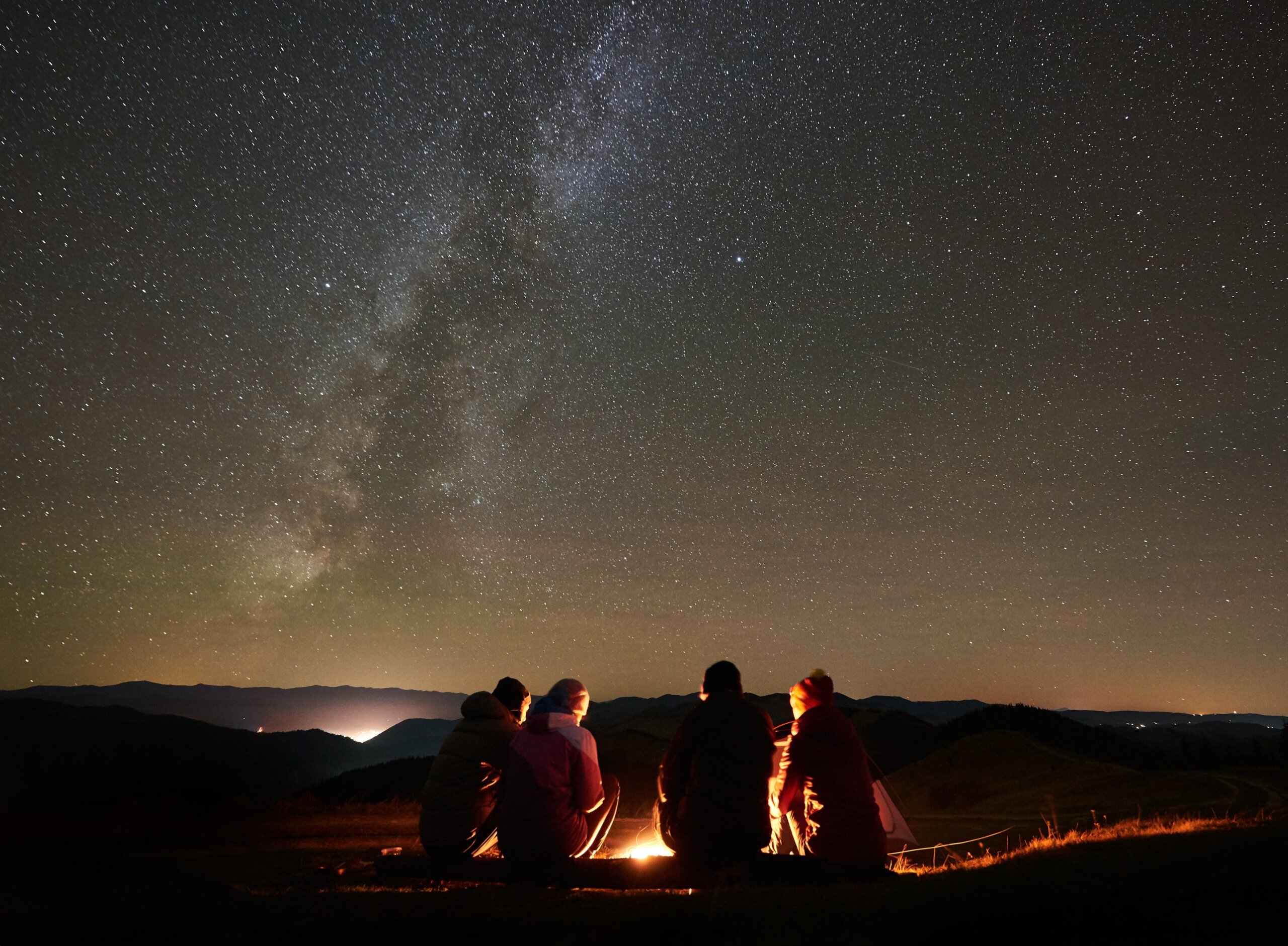 Night summer camping in the mountains. Back view group of four friends tourists sitting on a bench made of logs together around campfire under amazing night starry sky full of stars and Milky way.