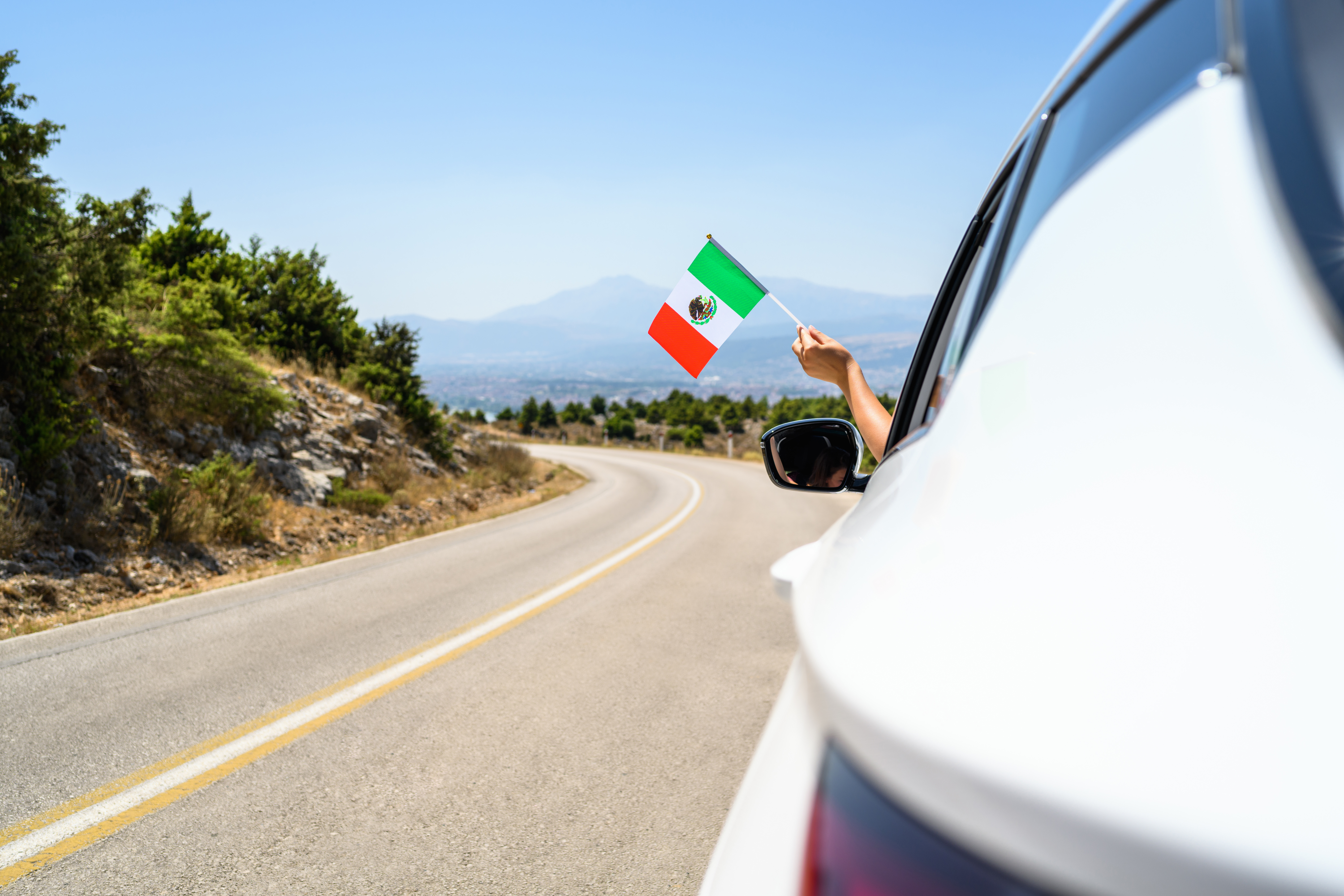 Woman holding Mexico flag from the open car window driving along the serpentine road in the mountains. Concept
