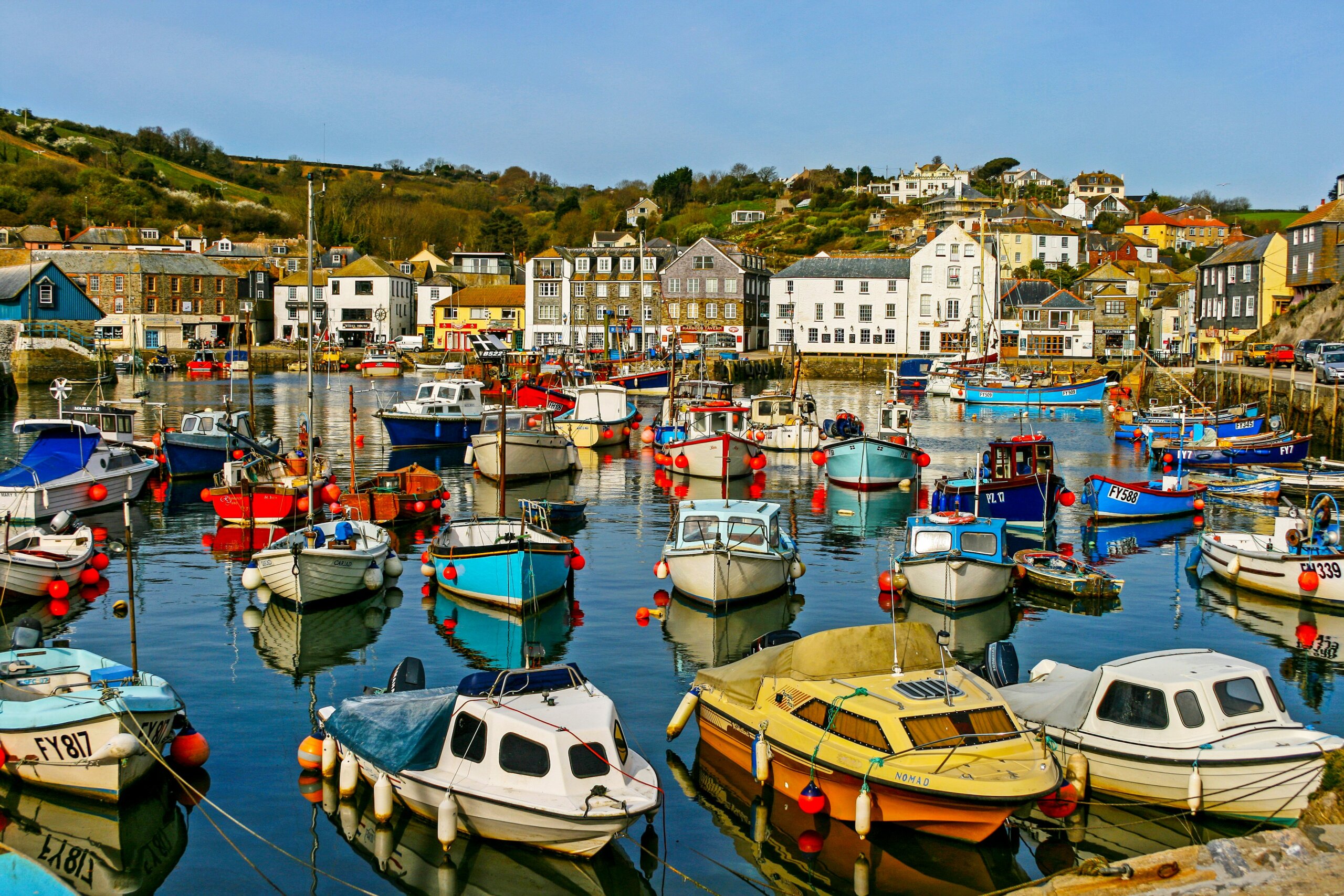 Colorful fishing boats docked in a calm Cornwall harbor with seaside cottages along the shore.