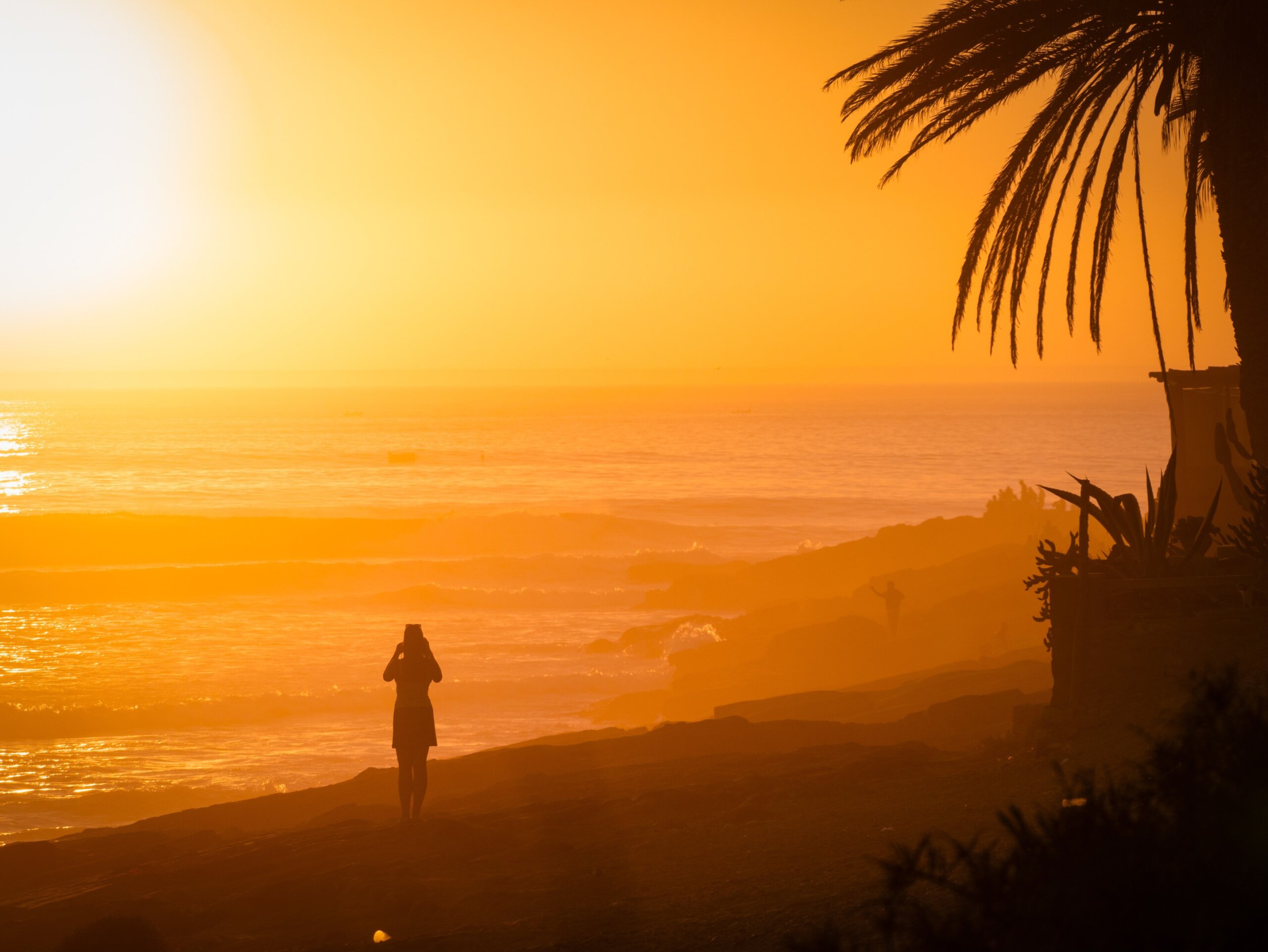 Golden sunset and female tourist holding a phone to capture the moment. Warm light illuminates breaking waves and rocky shoreline, showcasing stunning scenery of Moroccan coast
