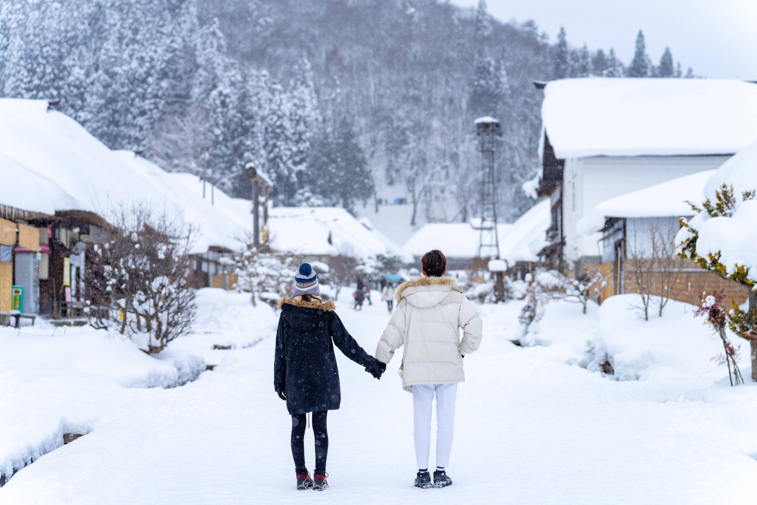 Happy young Asian couple in winter coat walking through the streets in snow. Man and woman traveler enjoy outdoor lifestyle travel countryside small town in Japan on winter holiday vacation.