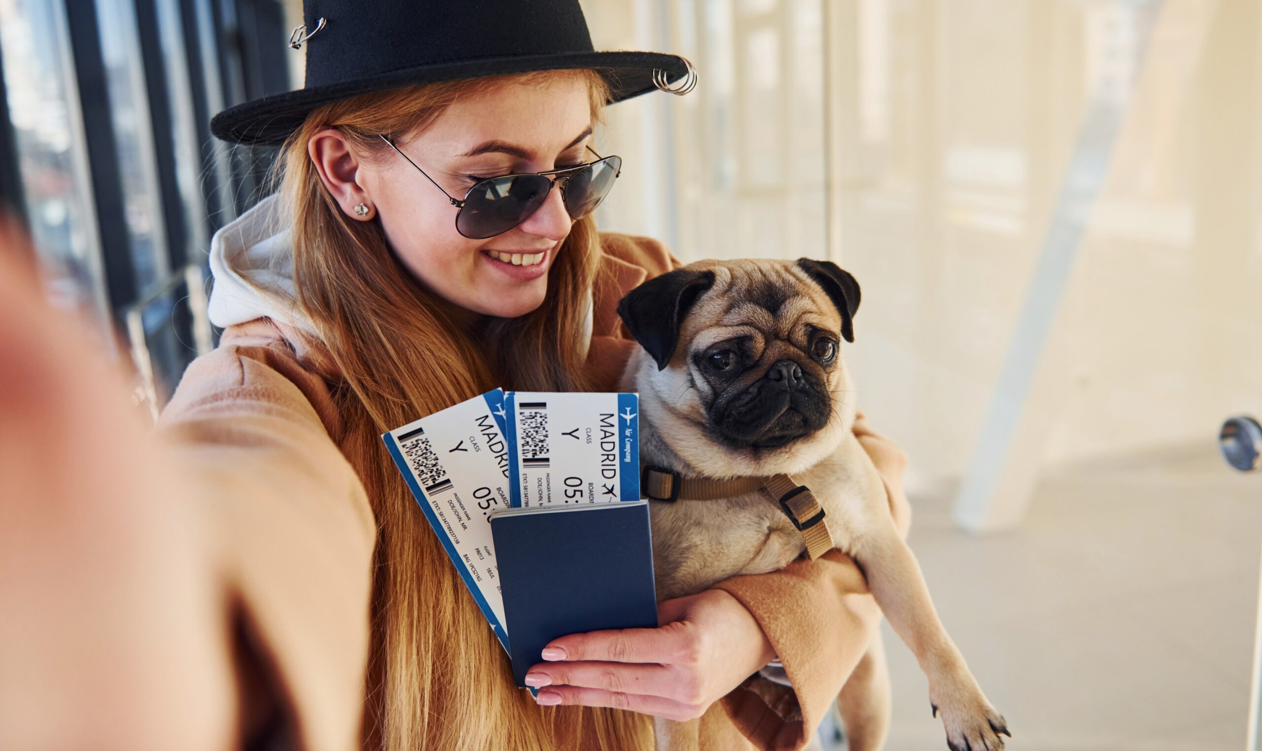 Young female passenger in warm clothes holding tickets and cute dog in hands in airport hall