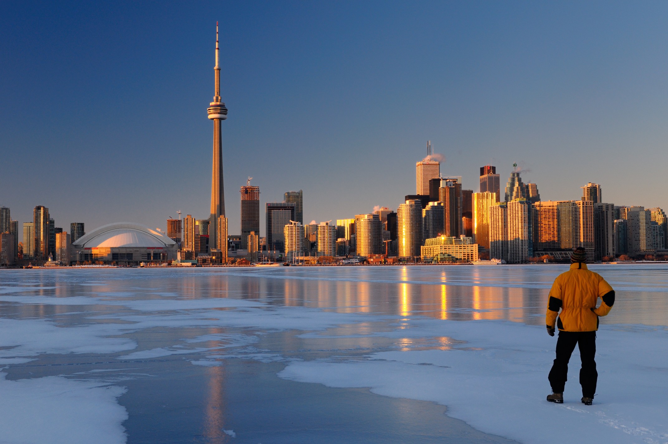 Man standing on frozen Lake Ontario ice looking at Toronto city skyline at sunset
