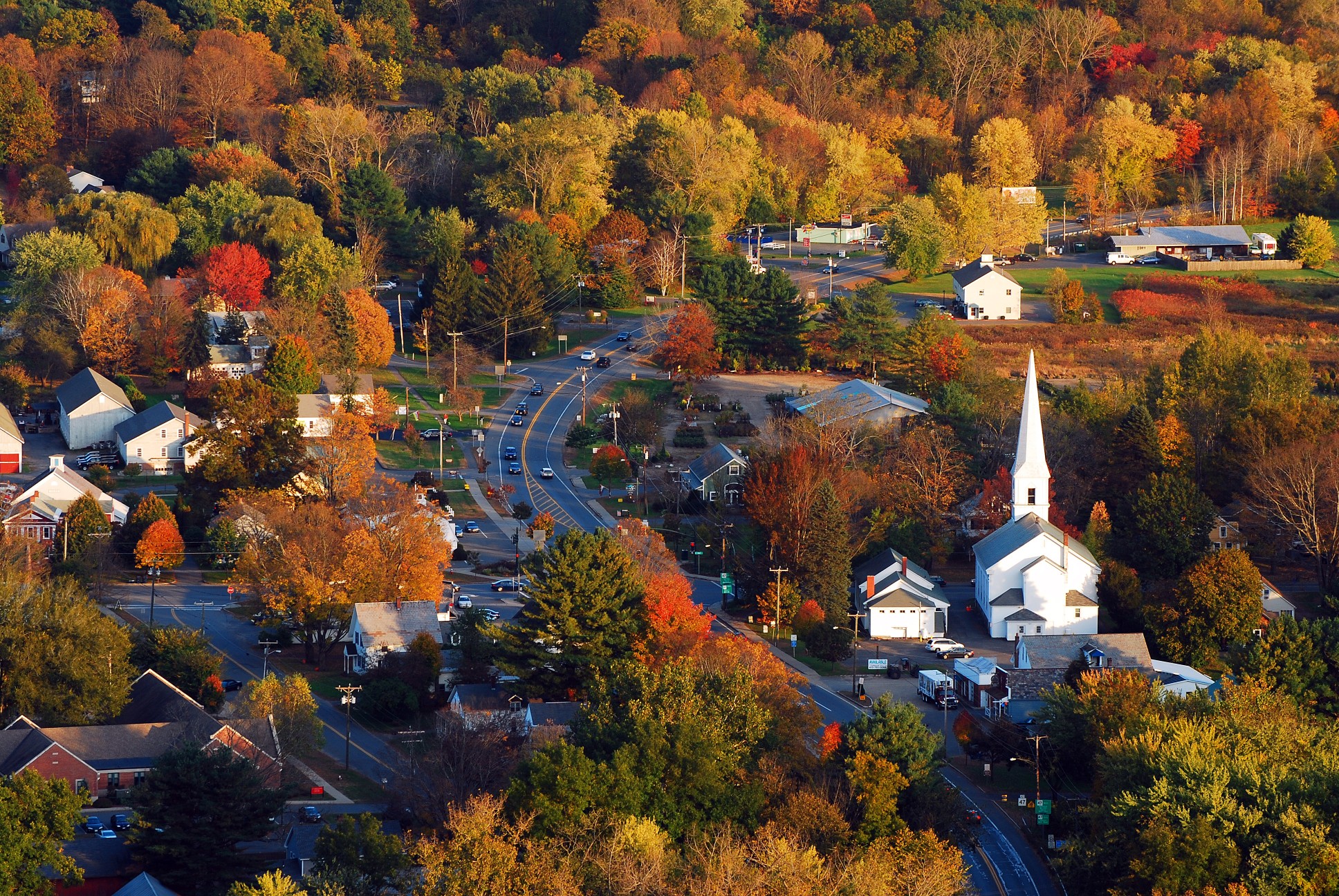 A classic New England town, with a white church with a large steeple, is surrounded by brilliant autumn foliage in an aerial view
