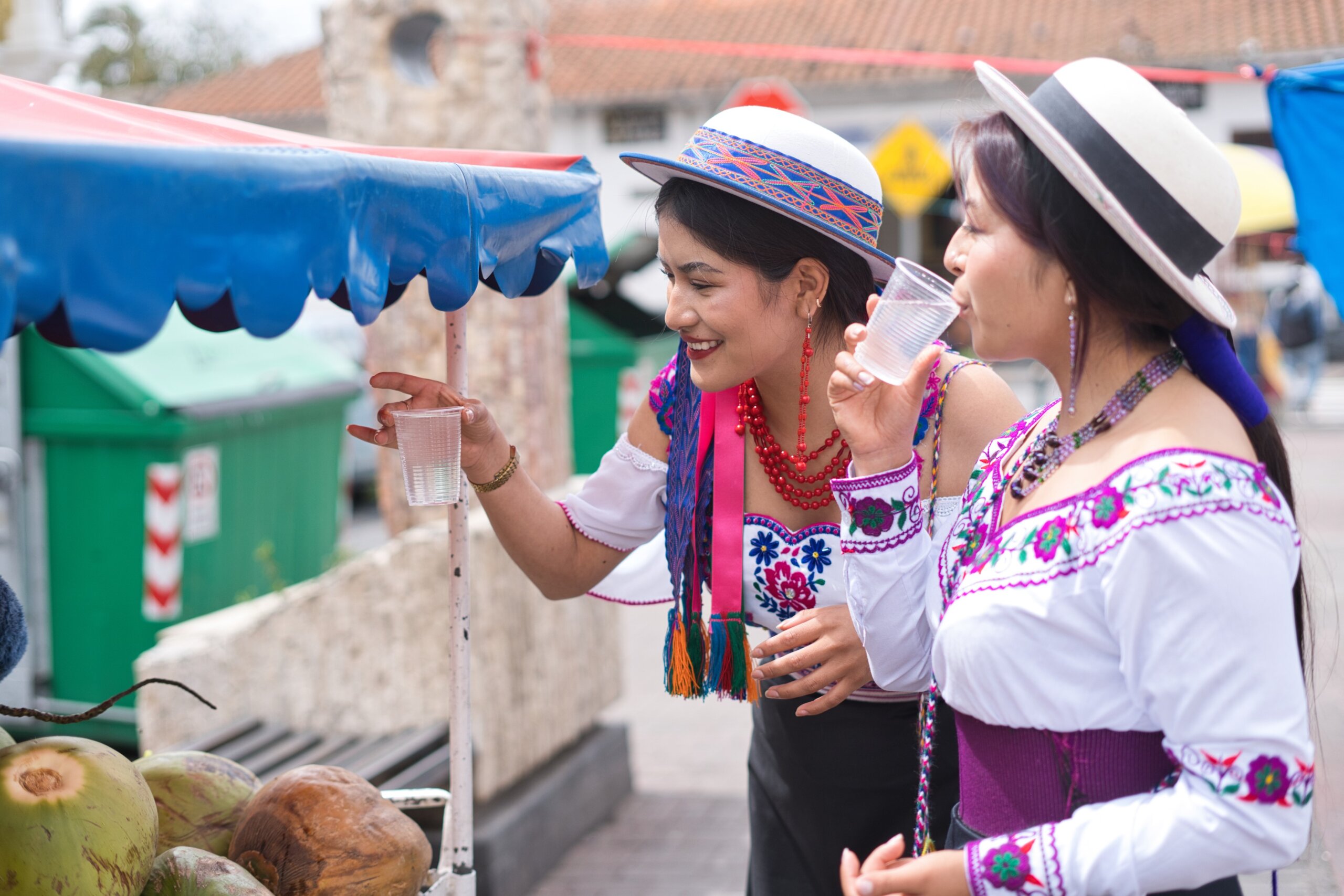 Two indigenous women in vibrant, traditional clothing are enjoying refreshing drinks from a street vendor
