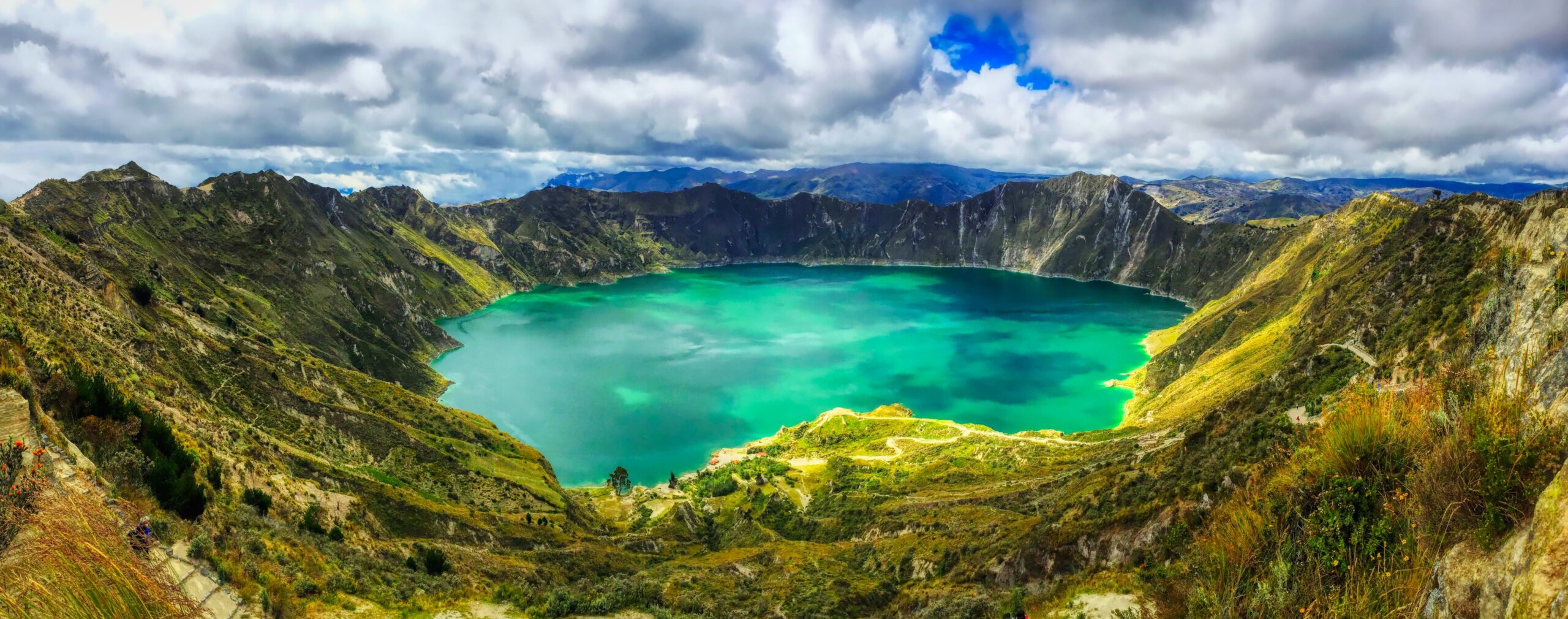 A bird's eye panoramic view of the bright green volcanic Quilotoa Lake in Ecuador with lots of white and grey clouds in a blue sky and green brush on the side of the caldera