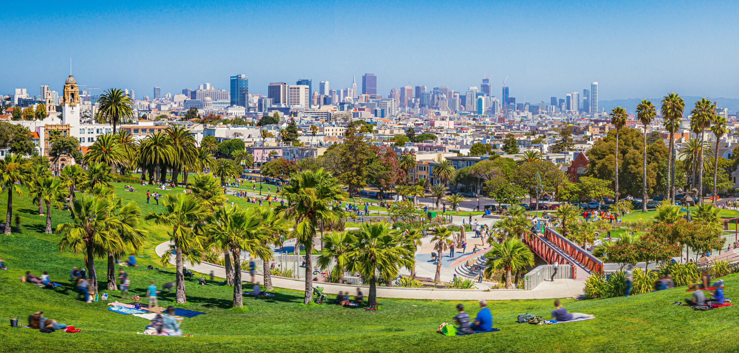 Panoramic view of local people enjoying the sunny summer weather at Mission Dolores Park on a beautiful day with clear blue sky with the skyline of San Francisco in the background, California, USA