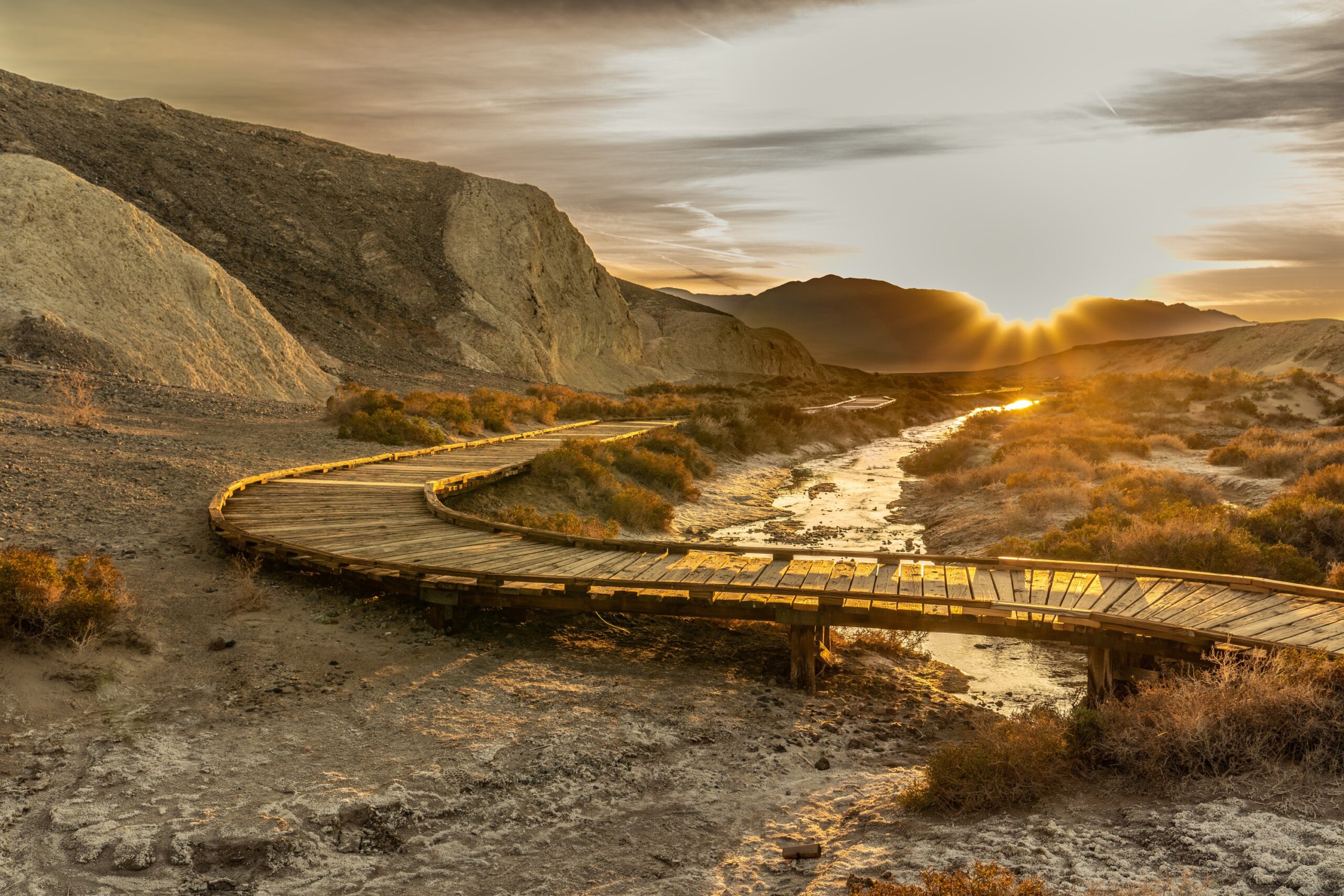 A wooden low level elevated walking path with planks winding through a wetland area with shrubs and a creek flowing in the center. The setting sun is shining over distance mountains.