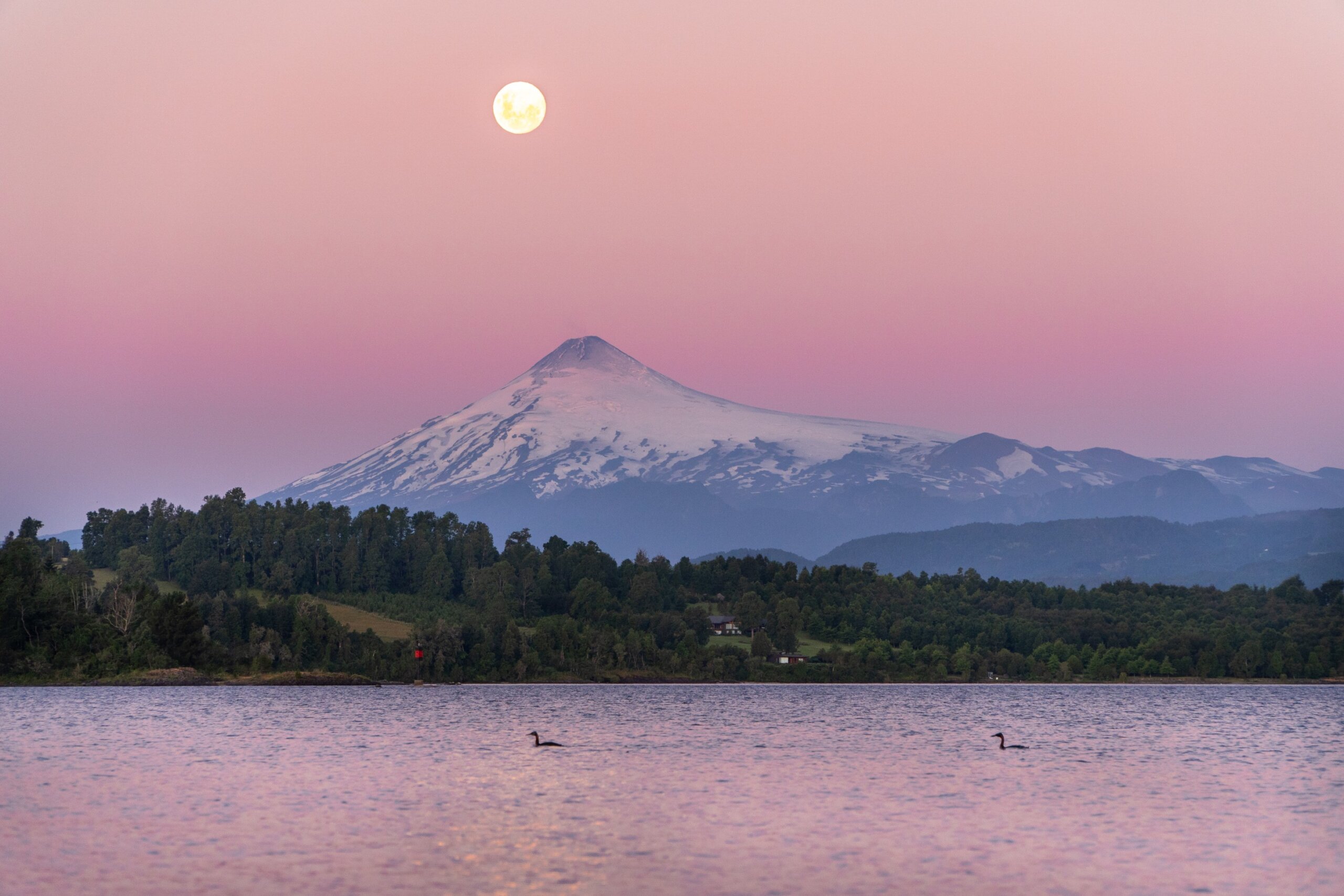 view of the Villarrica volcano reflected in the lake, with the full moon shining during a stunning sunset