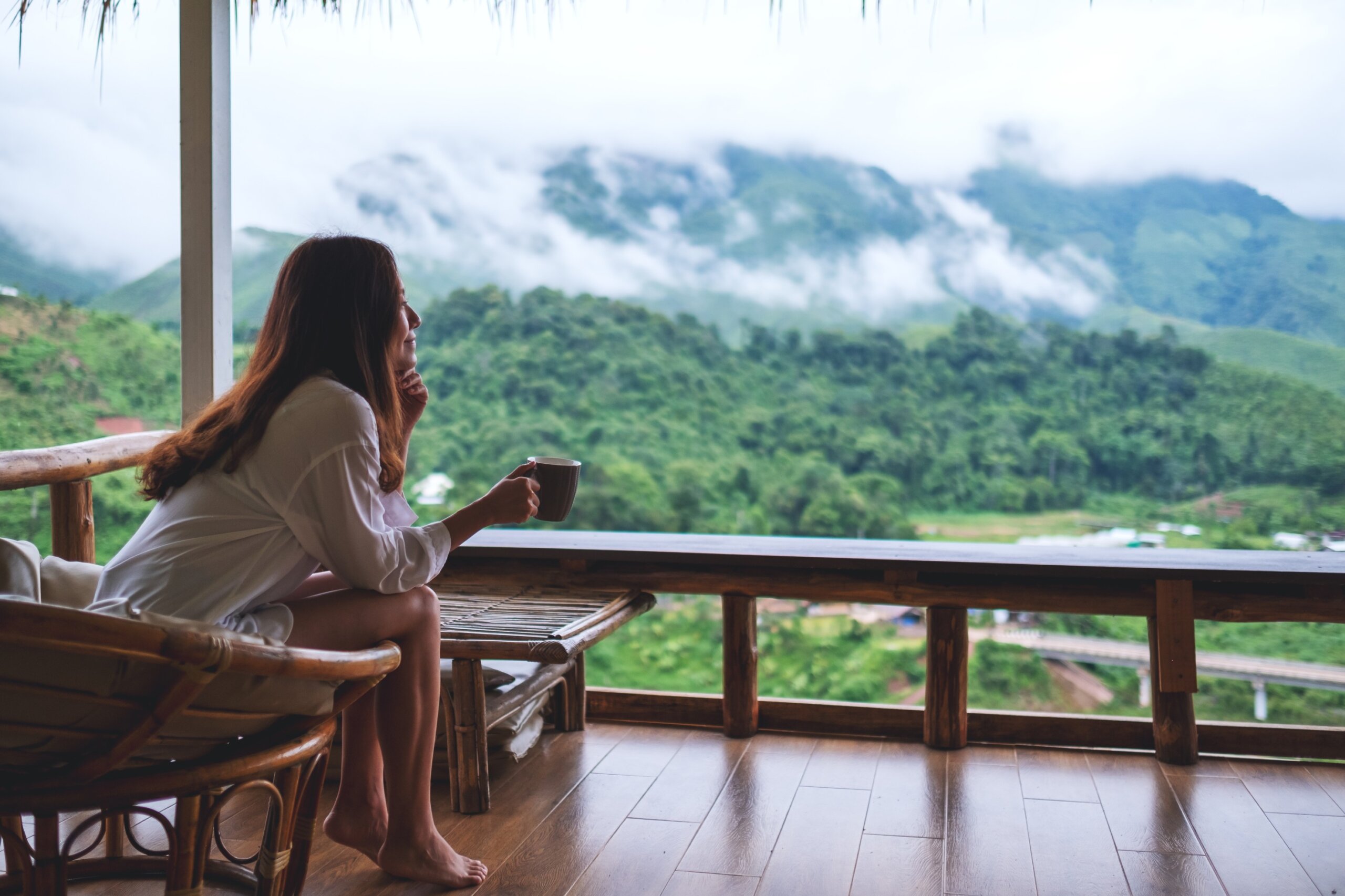 Portrait image of a beautiful asian woman holding and drinking hot coffee on balcony , looking at mountains and green nature