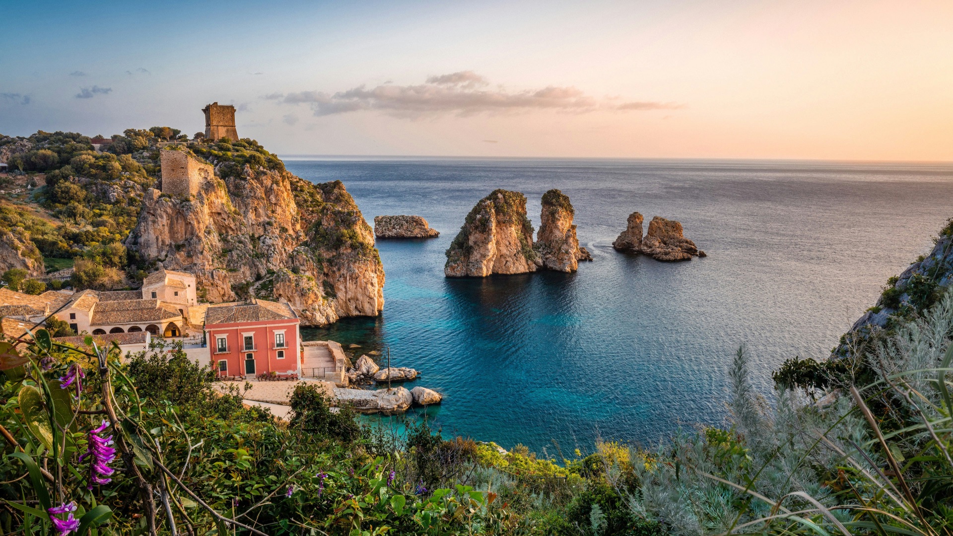 Dramatic rocky sea stacks and a cliffside stone village overlooking turquoise Mediterranean water at sunset with wildflowers in the foreground
