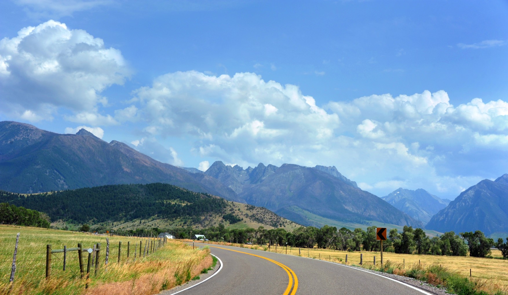 Two-lane highway curving toward a dramatic mountain range with golden fields and wooden fences under a bright blue sky in Big Sky Montana
