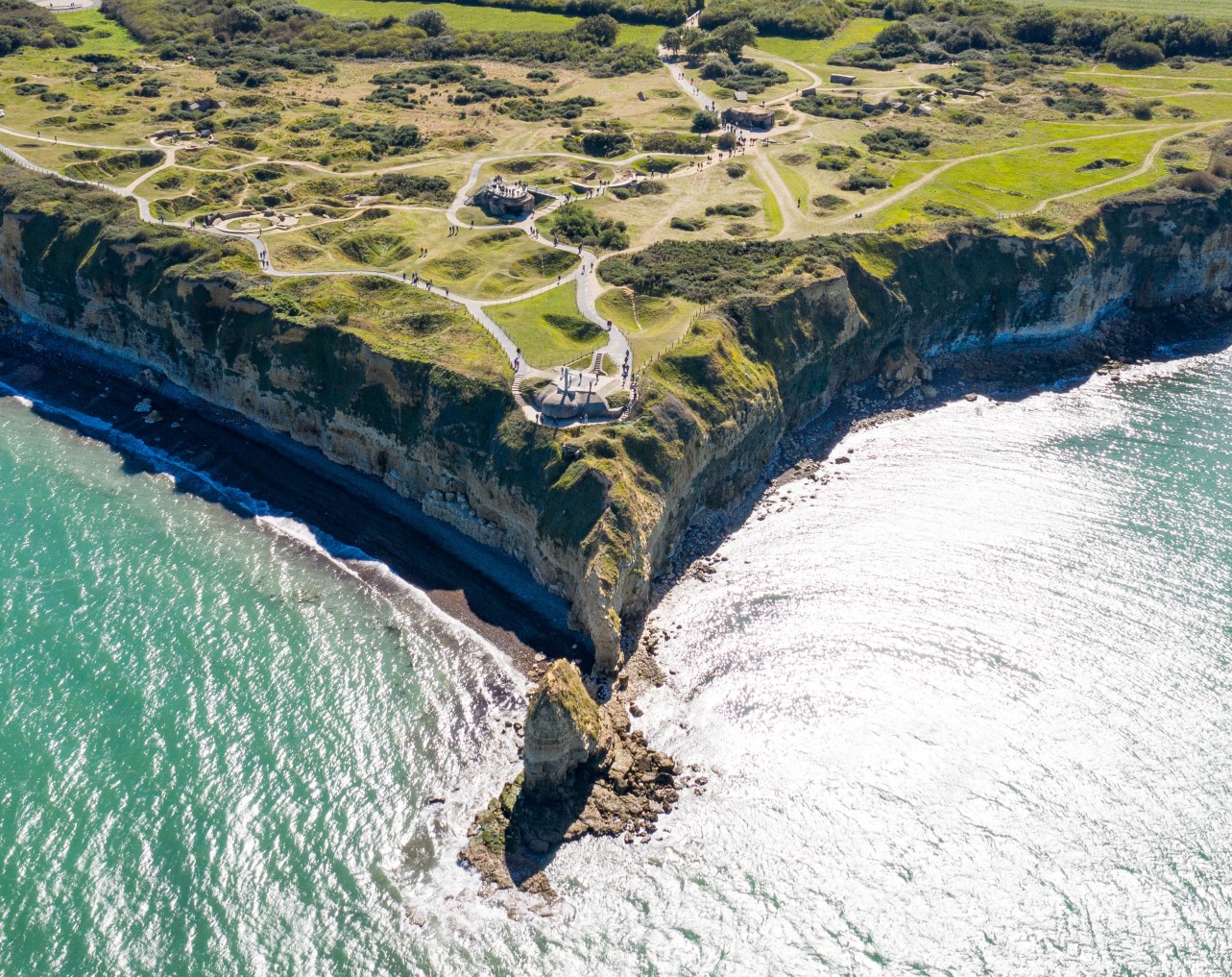 Aerial view of Pointe du Hoc Normandy cliffs with WWII concrete bunkers, bomb craters, and walking paths above turquoise coastal waters